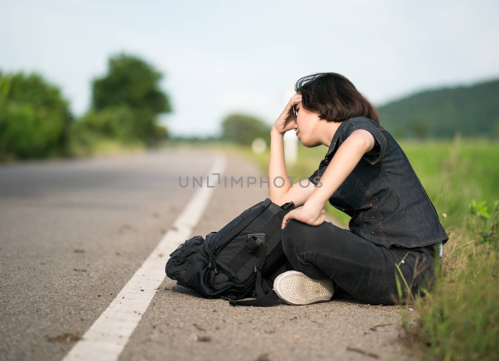 Young asian woman short hair and wearing sunglasses sit with backpack hitchhiking along a road wait for help in country road Thailand