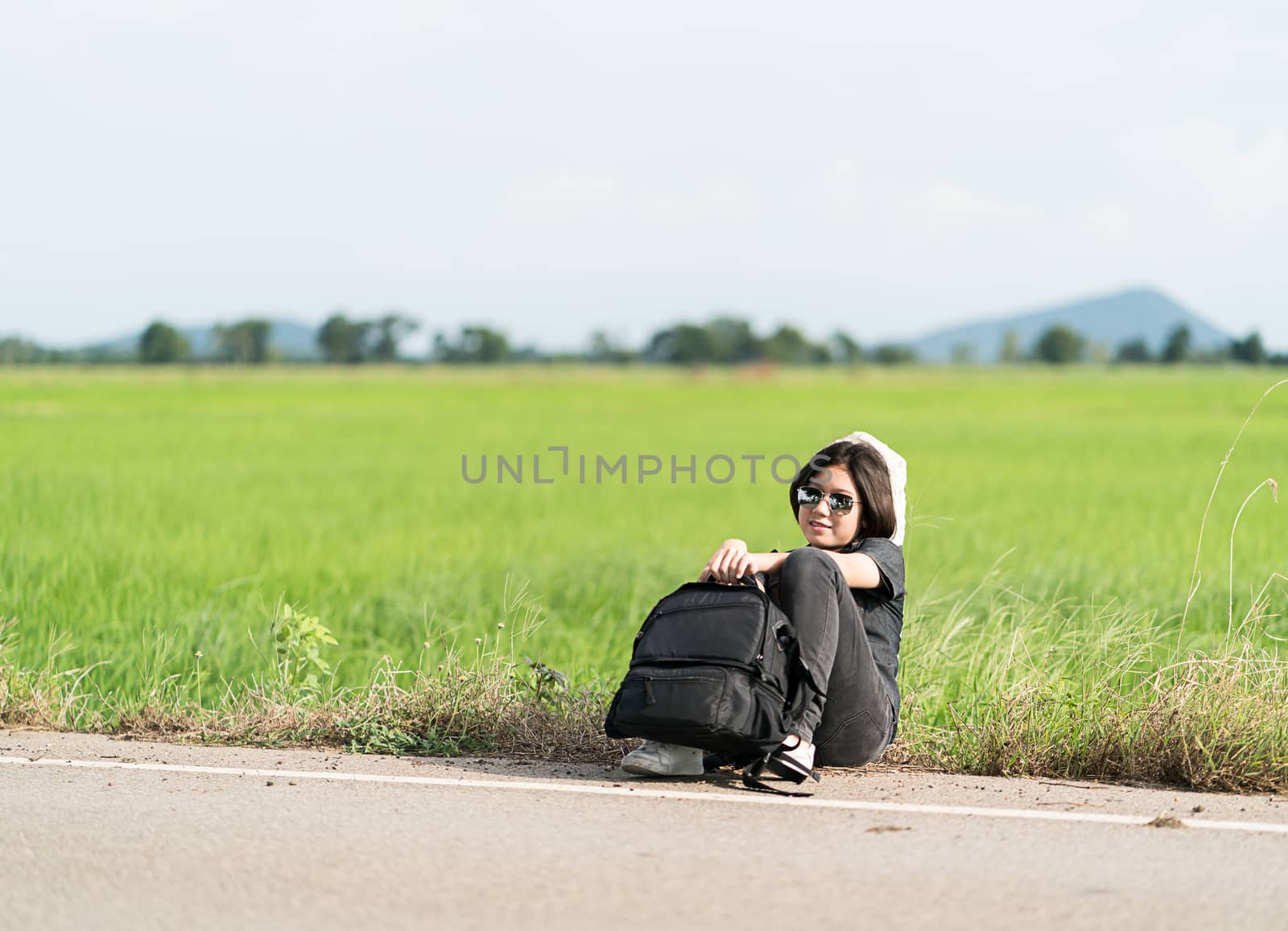 Young asian woman short hair and wearing sunglasses sit with backpack hitchhiking along a road wait for help in countryside Thailand