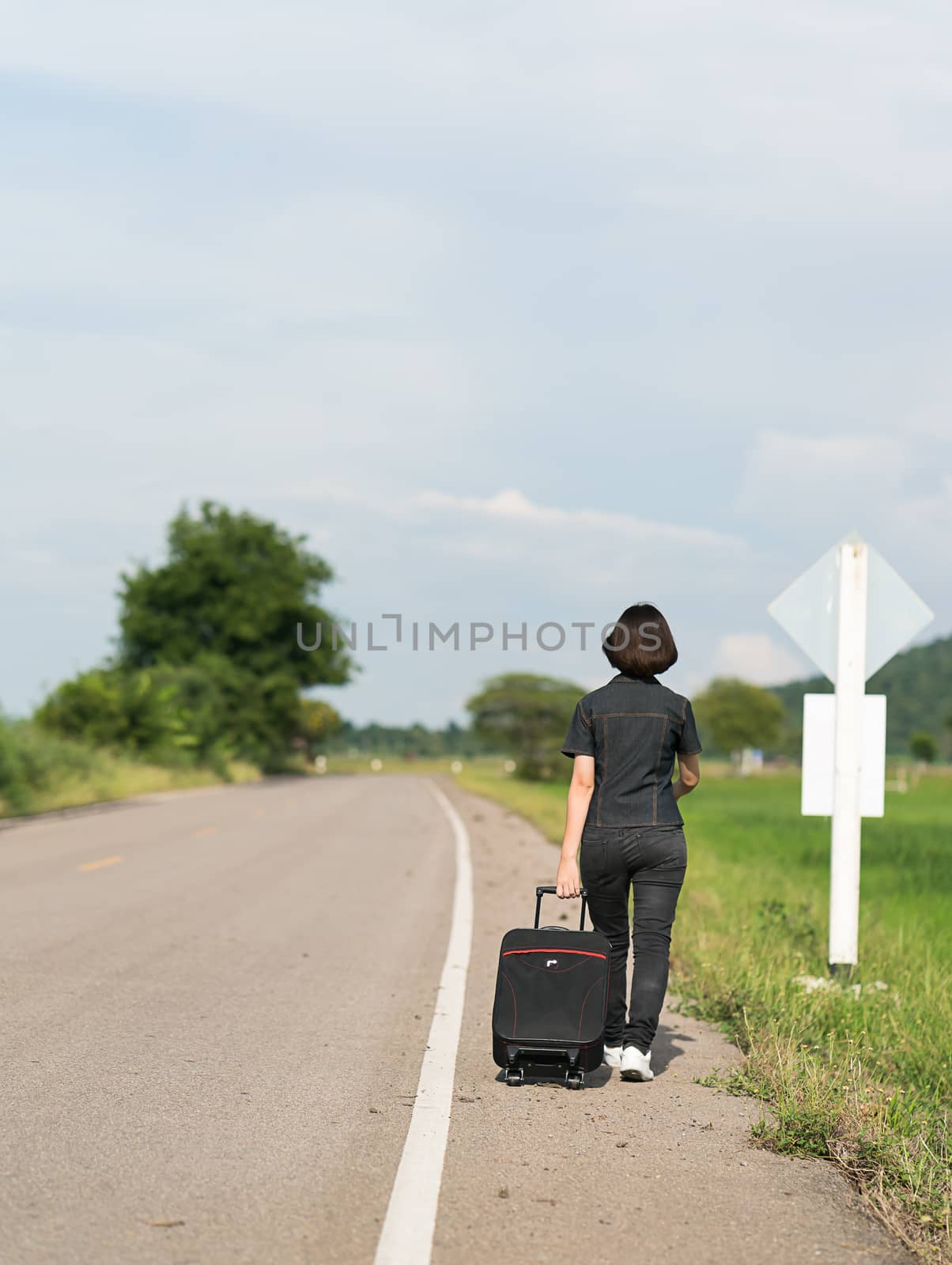 Young asian woman short hair and wearing sunglasses with luggage hitchhiking along a road in countryside Thailand