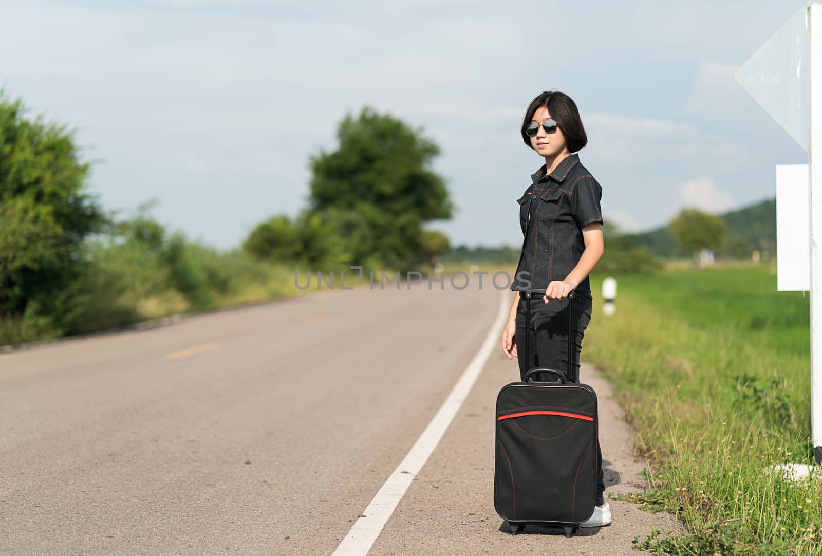 Young asian woman short hair and wearing sunglasses with luggage hitchhiking along a road in countryside Thailand