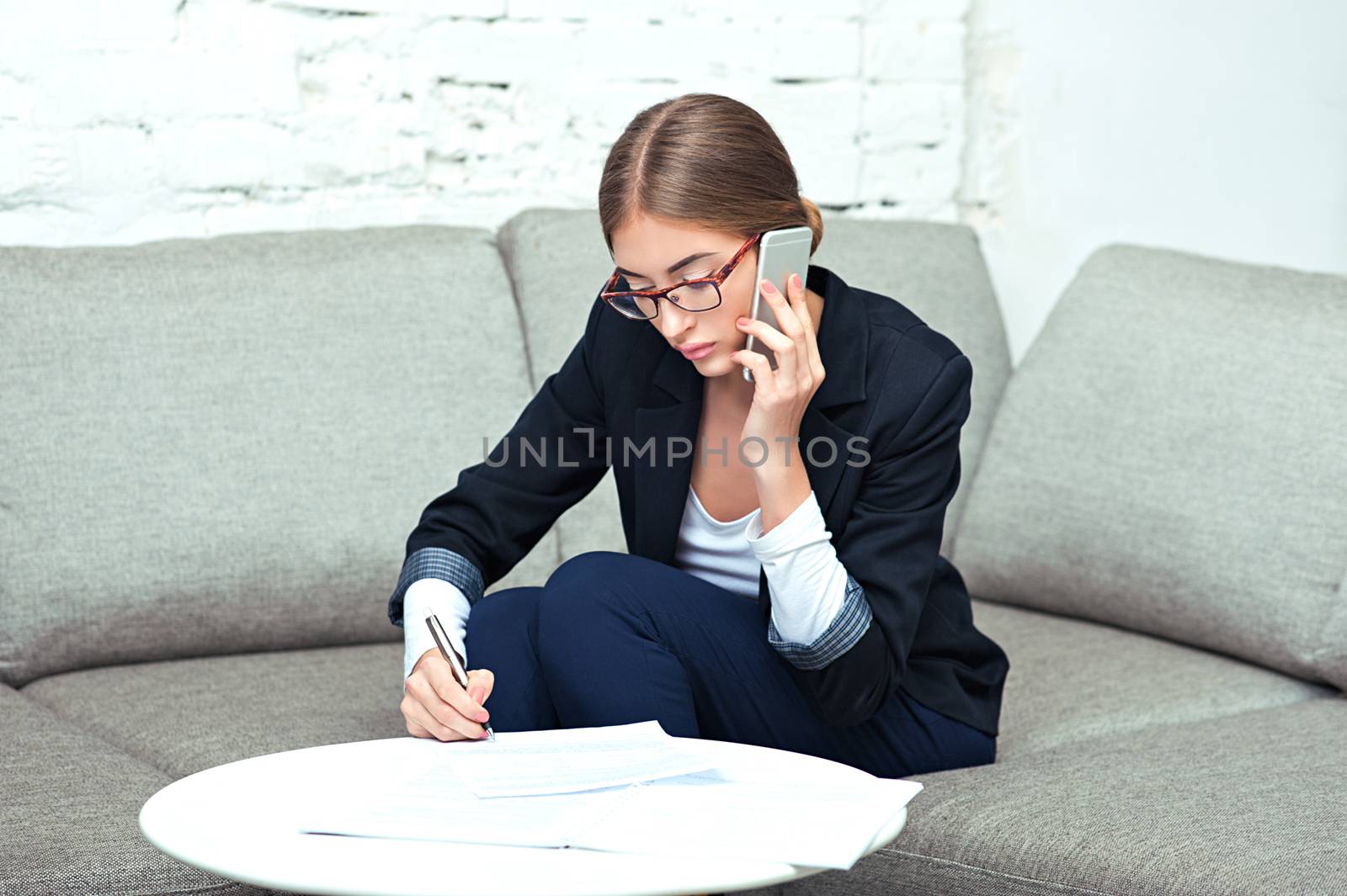 Beautiful young business woman sitting on sofa at office or at home, calling on phone and working with documents