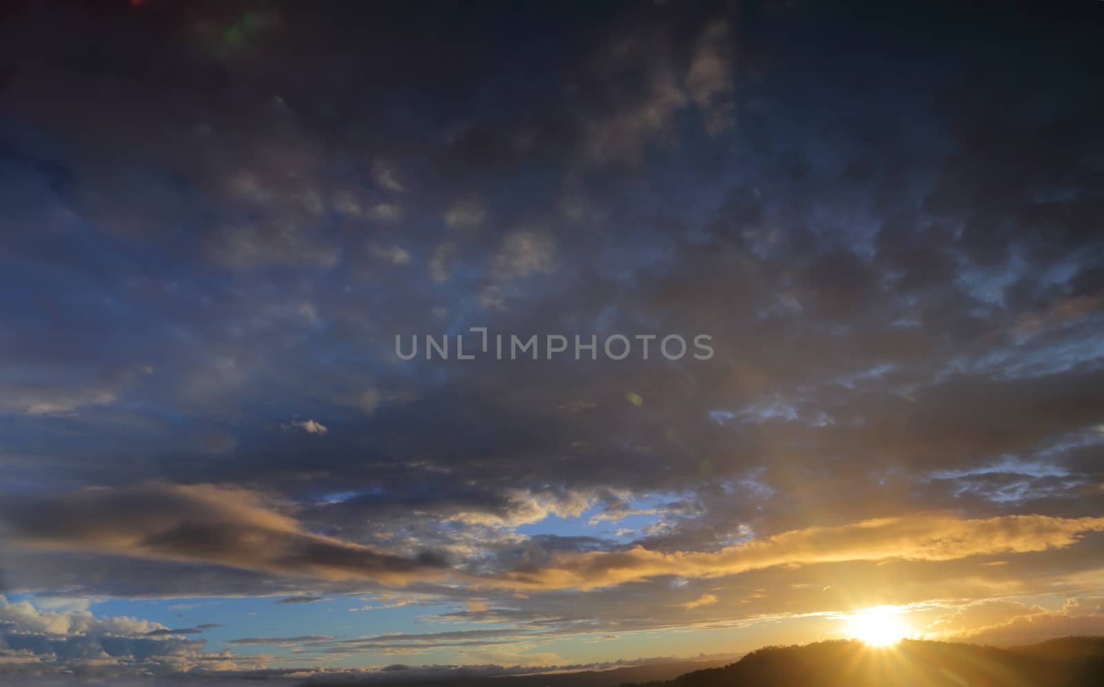 Sun setting sunburst behind mountain with blue sky and clouds tinged with colour.  The image shows primarily sky