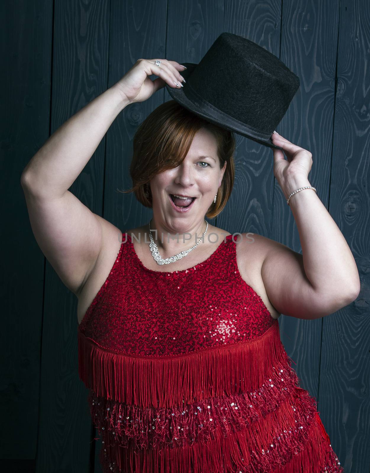 forty something brunette woman wearing a red sparkling dress dancing with a hat in front of blue wood panel