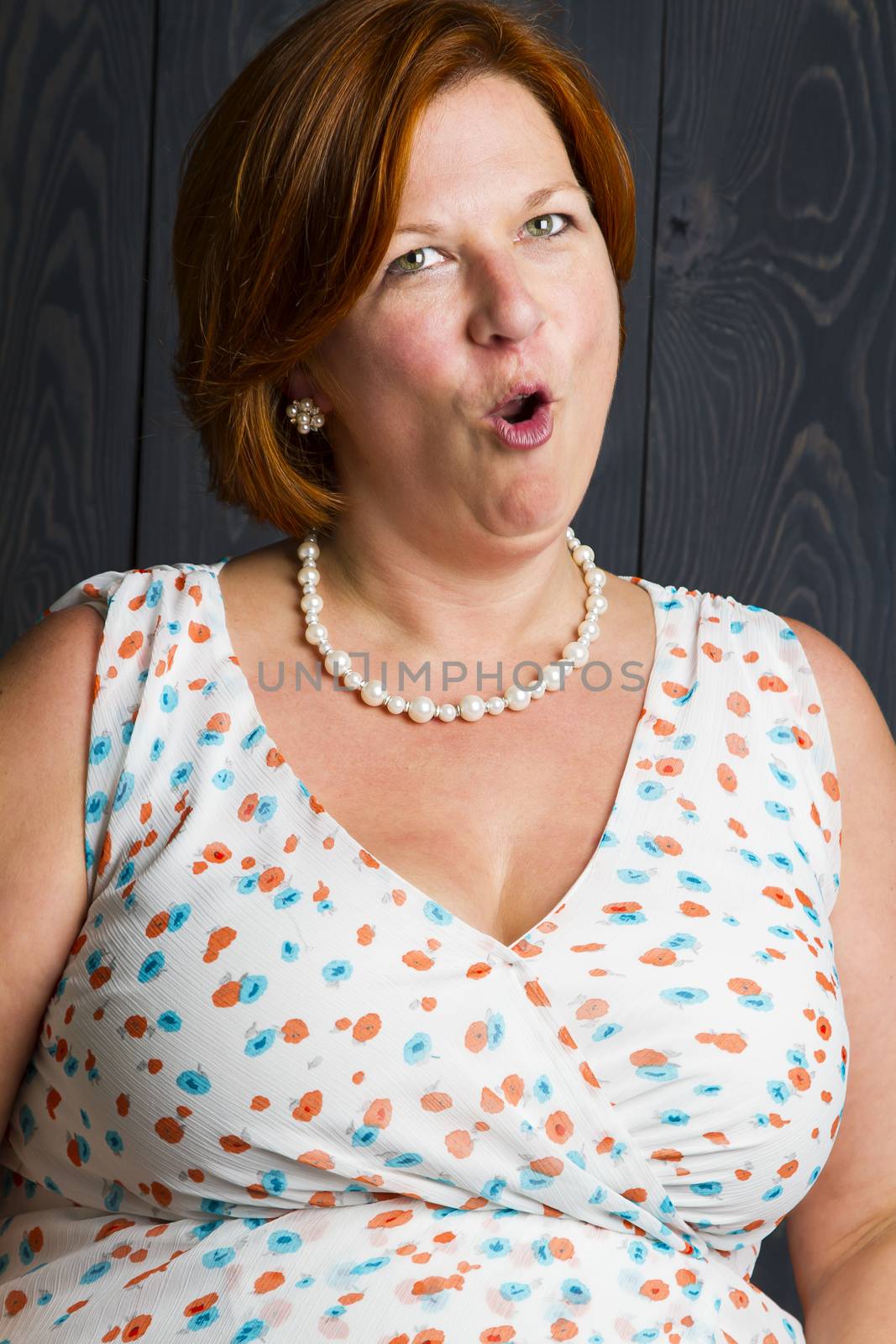 forty something woman wearing a sundress in front of a blue stain background, doing a surprise "O' face