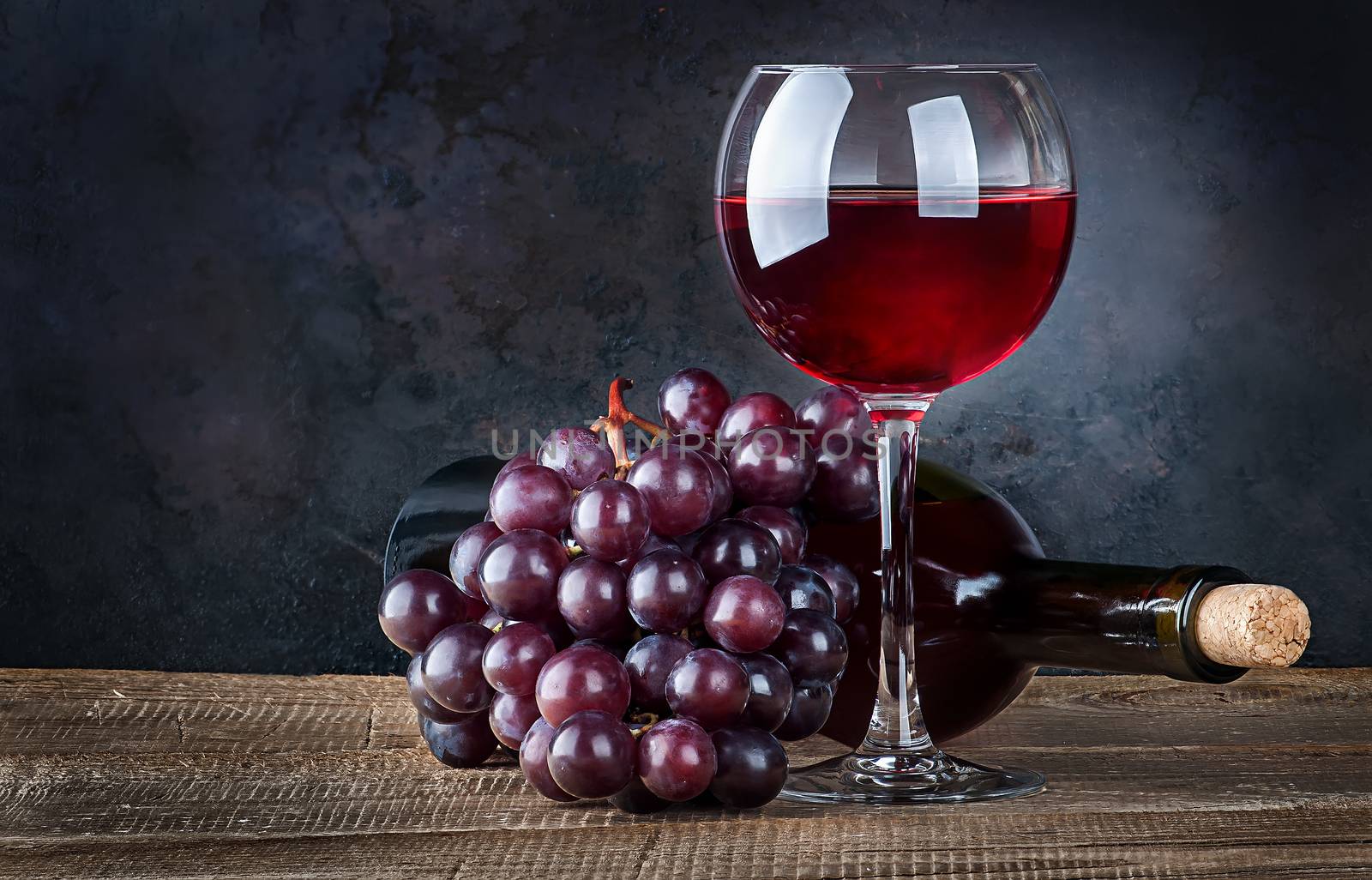 Glass of red wine with grapes and bottle on a wooden table. The bottle lies. Dark background.