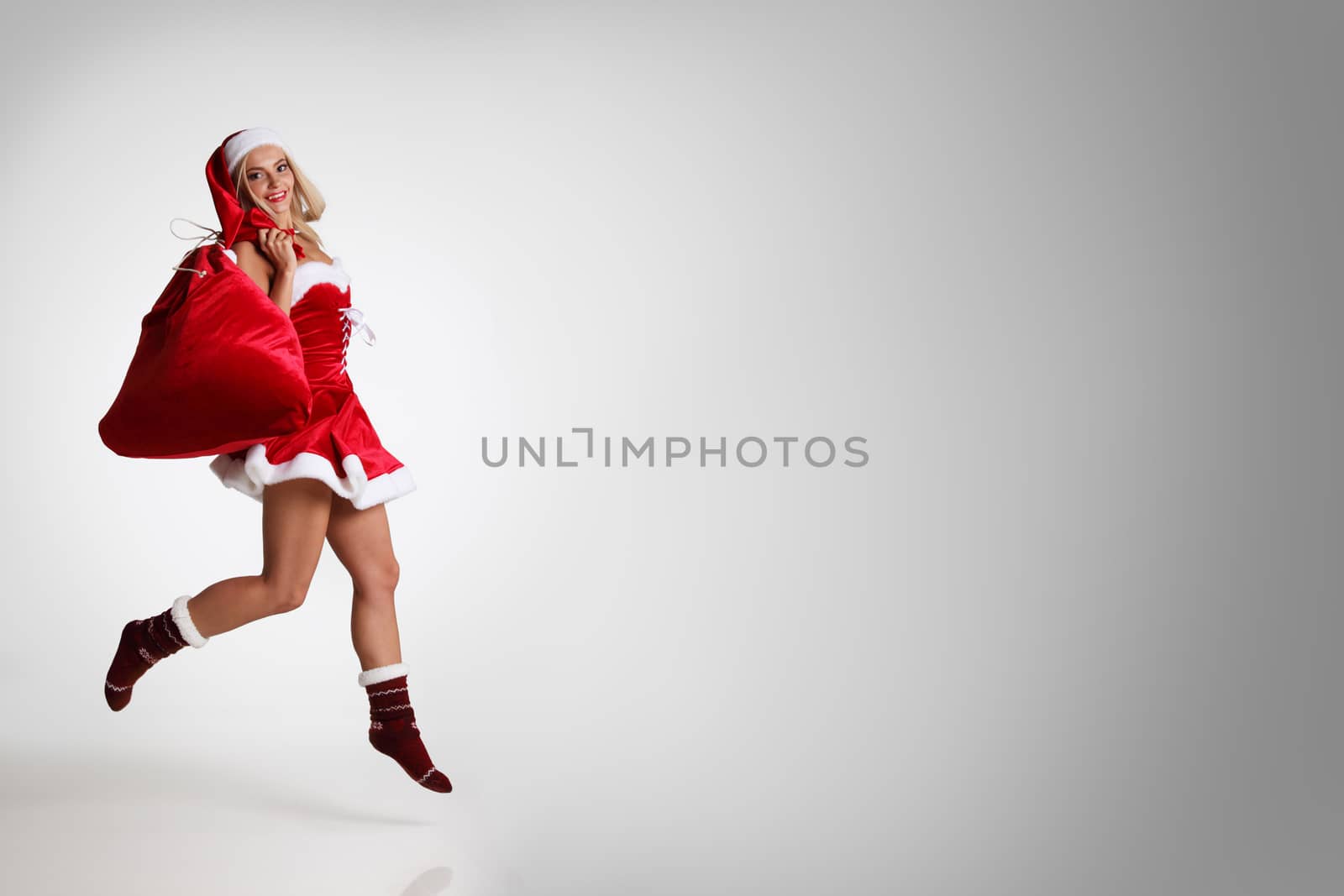 Sexy young Santa woman jumping with bag of christmas gifts, gray background with copy space