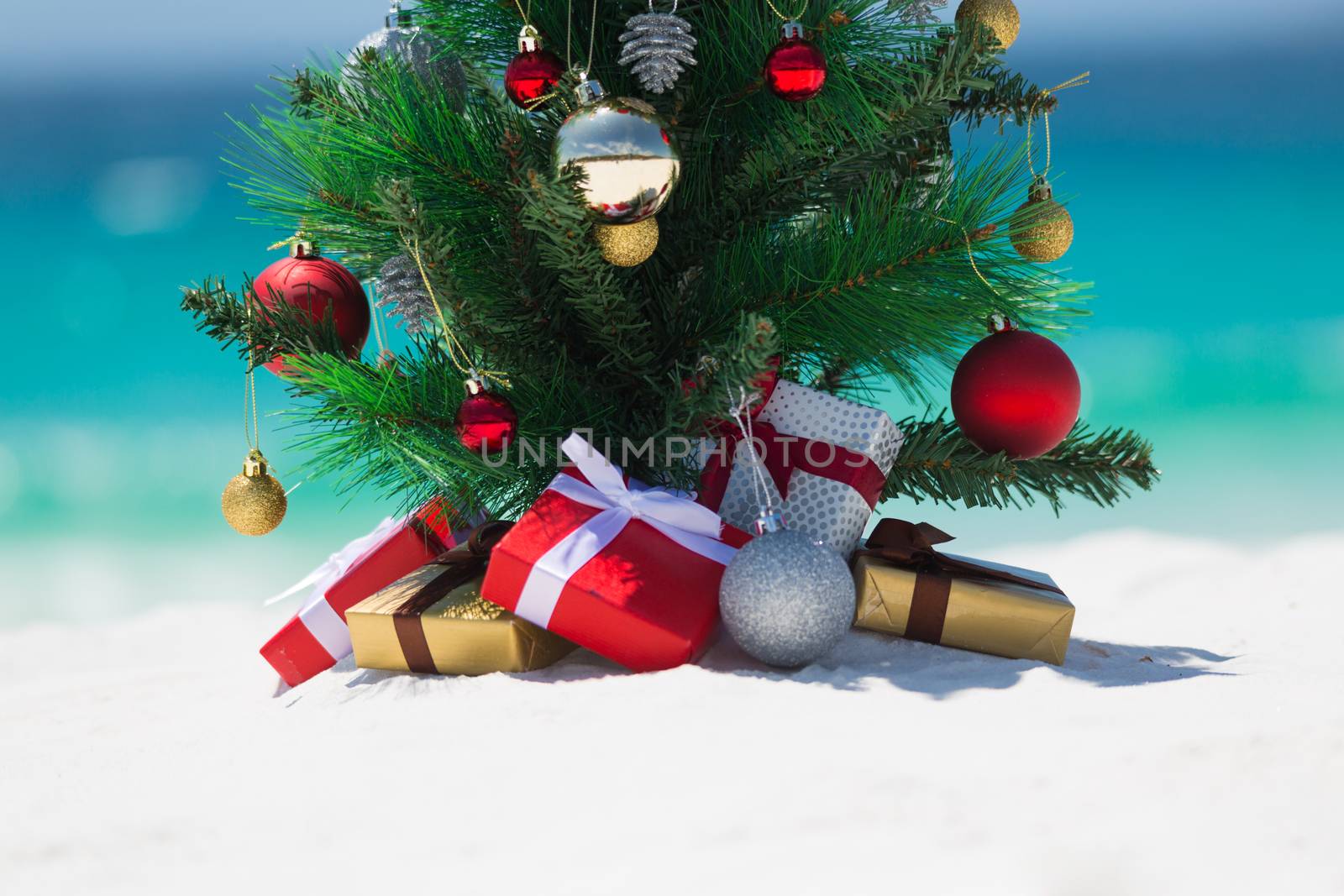 Christmas in some countries is spent in the summer months.  A beautiful christmas tree sits on a white sandy beach under the summer sun.  There are wrapped gifts underneath it.  Space beneath for some copy or message