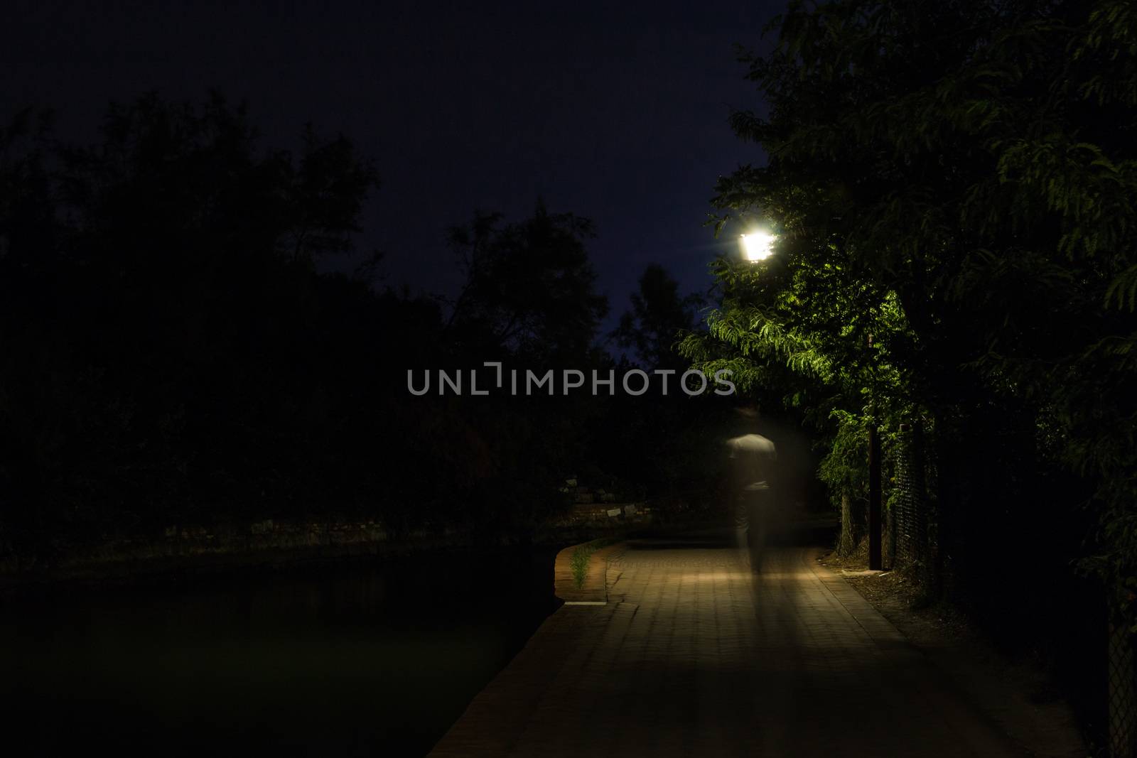 Double exposure night scene of person walking dark street illuminated with streetlights. The receding male silhouettes on the road in the park. Human figure in motion blur going along the city river. Long shutter.