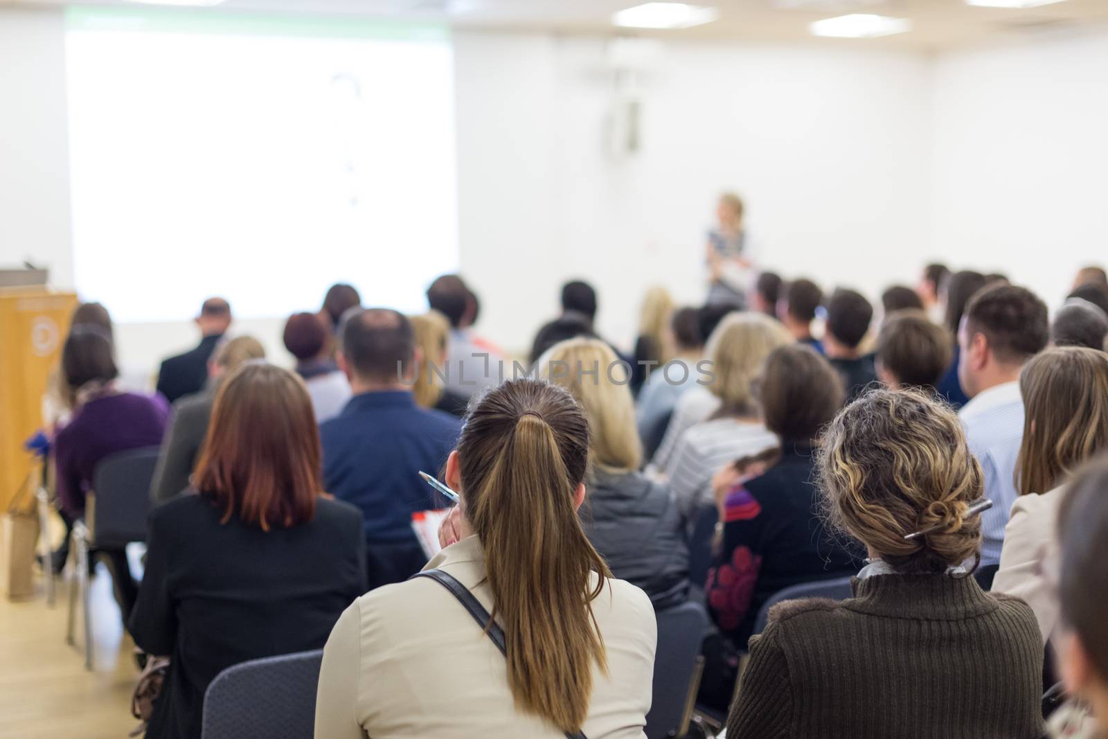 Business and entrepreneurship symposium. Female speaker giving a talk at business meeting. Audience in conference hall. Rear view of unrecognized participant in audience. Copy space on whitescreen.