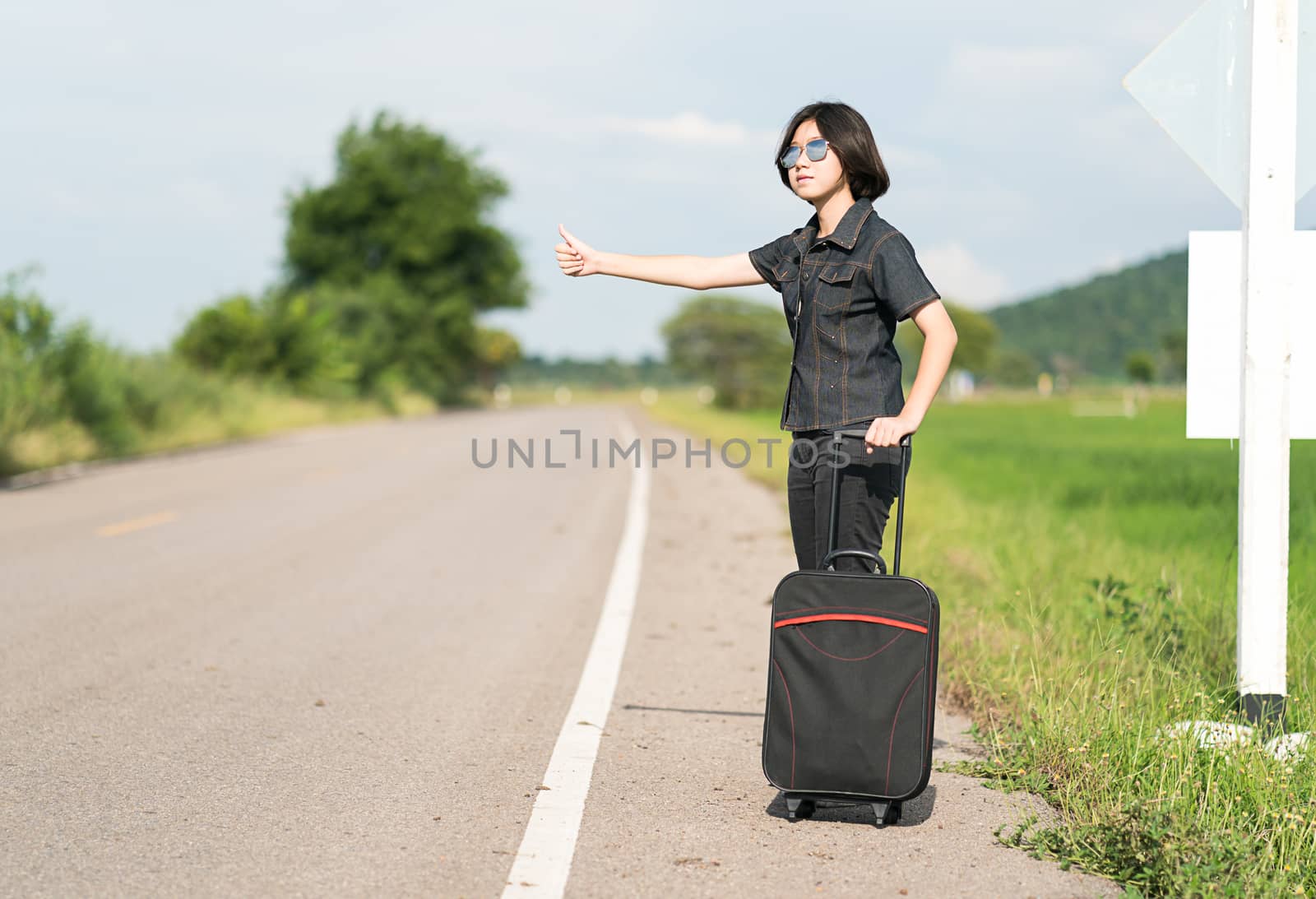 Young asian woman short hair and wearing sunglasses with luggage hitchhiking along a road and thumbs up in country road Thailand