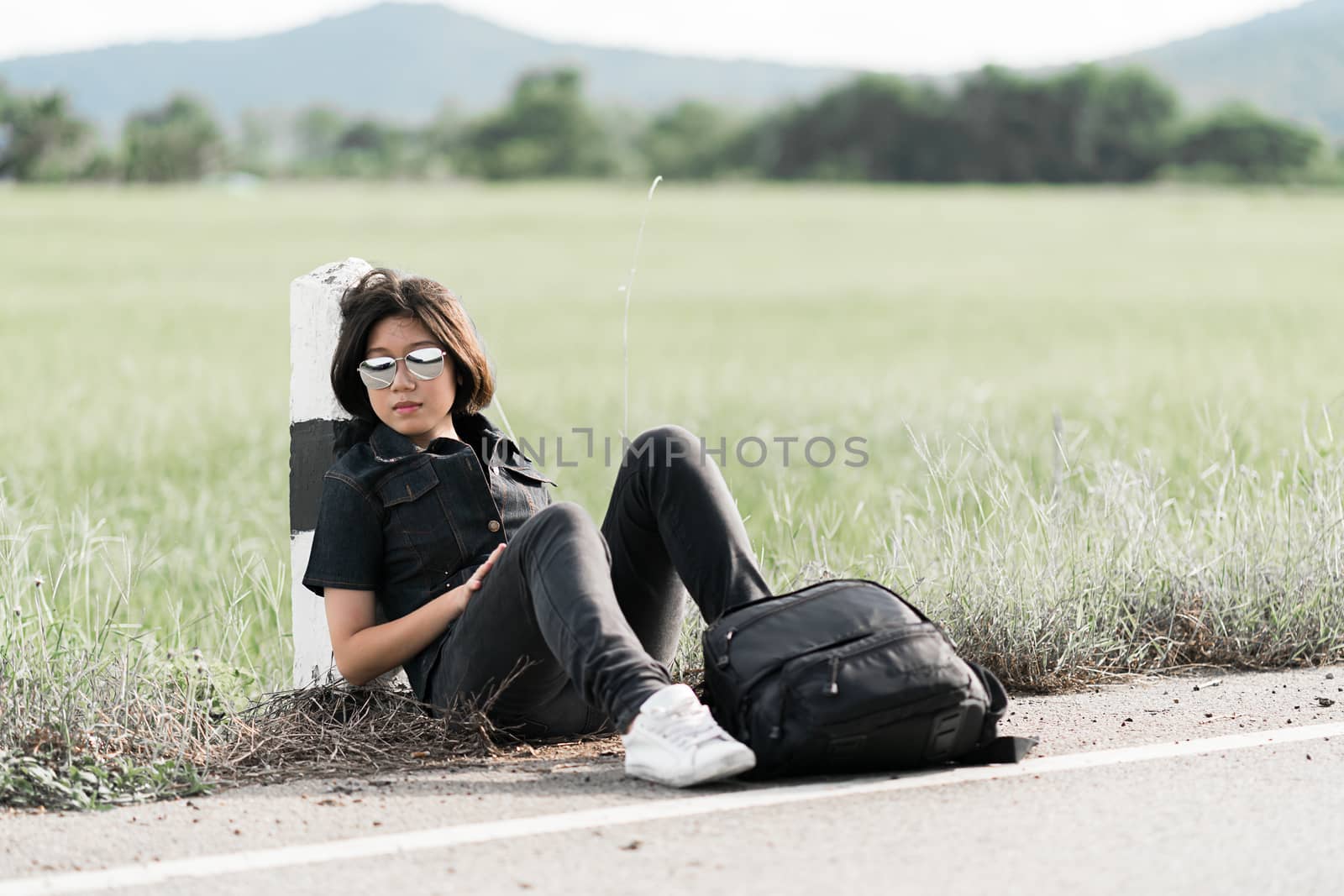 Young asian woman short hair and wearing sunglasses sit with backpack hitchhiking along a road wait for help in countryside Thailand