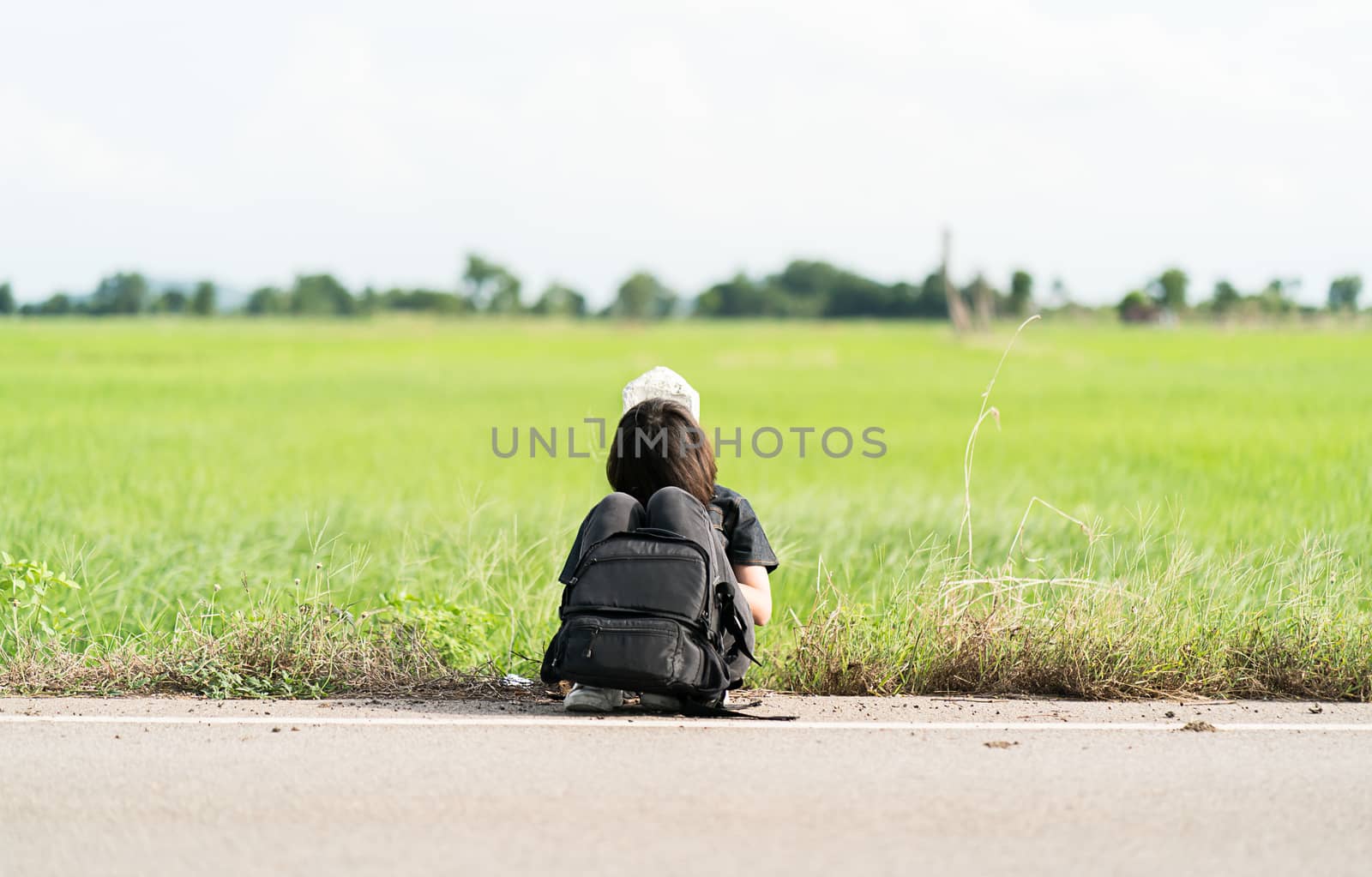 Young asian woman short hair and wearing sunglasses sit with backpack hitchhiking along a road wait for help in countryside Thailand