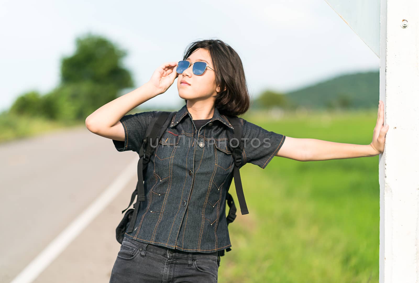 Young asian woman short hair and wearing sunglasses with backpack hitchhiking along a road in countryside Thailand