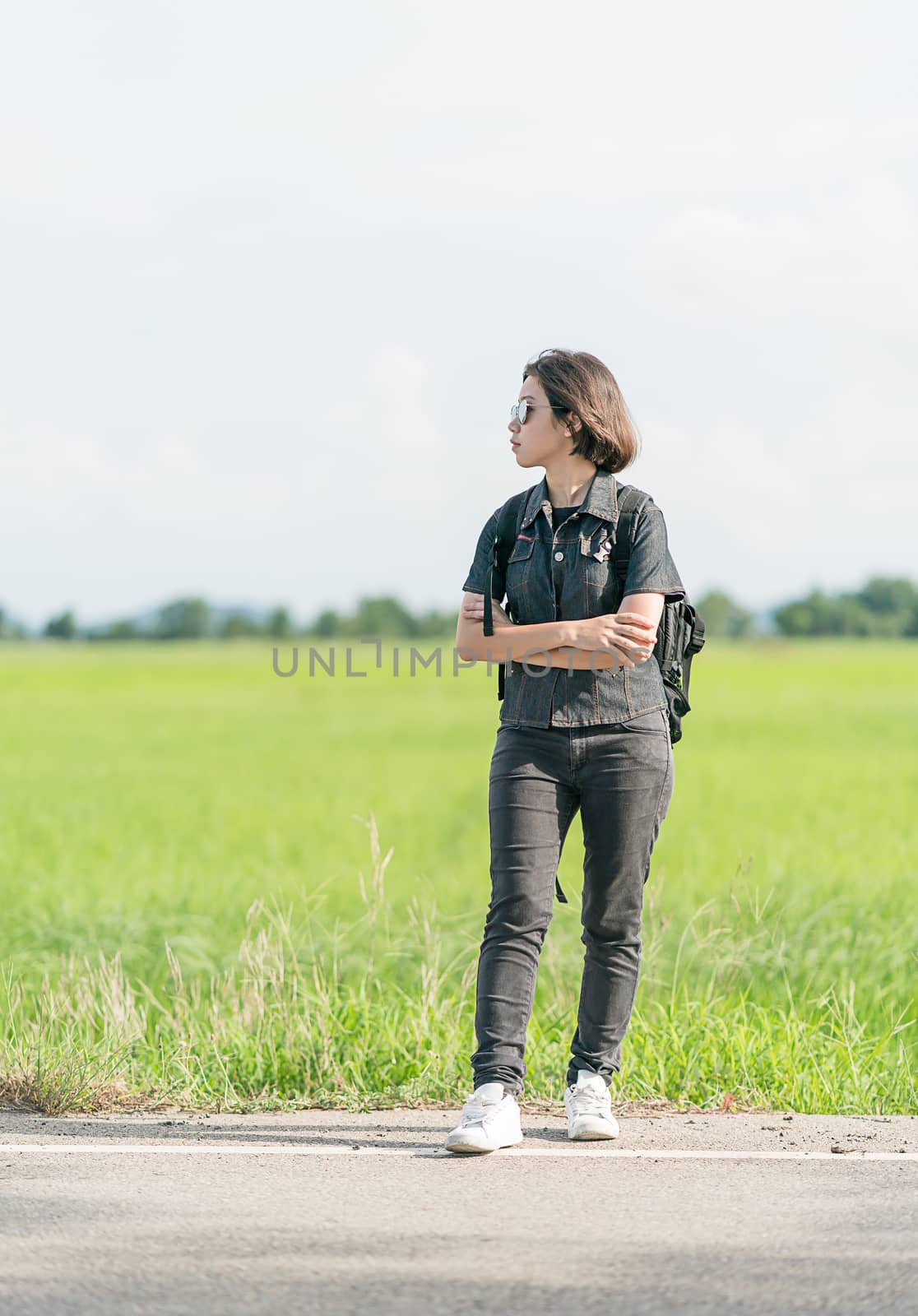 Young asian woman short hair and wearing sunglasses with backpack hitchhiking along a road in countryside Thailand