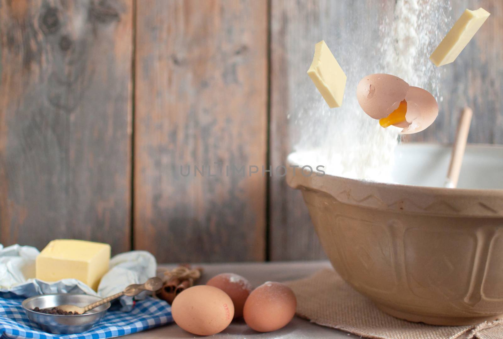 Eggs, flour and butter falling into a mixing bowl over a wooden background