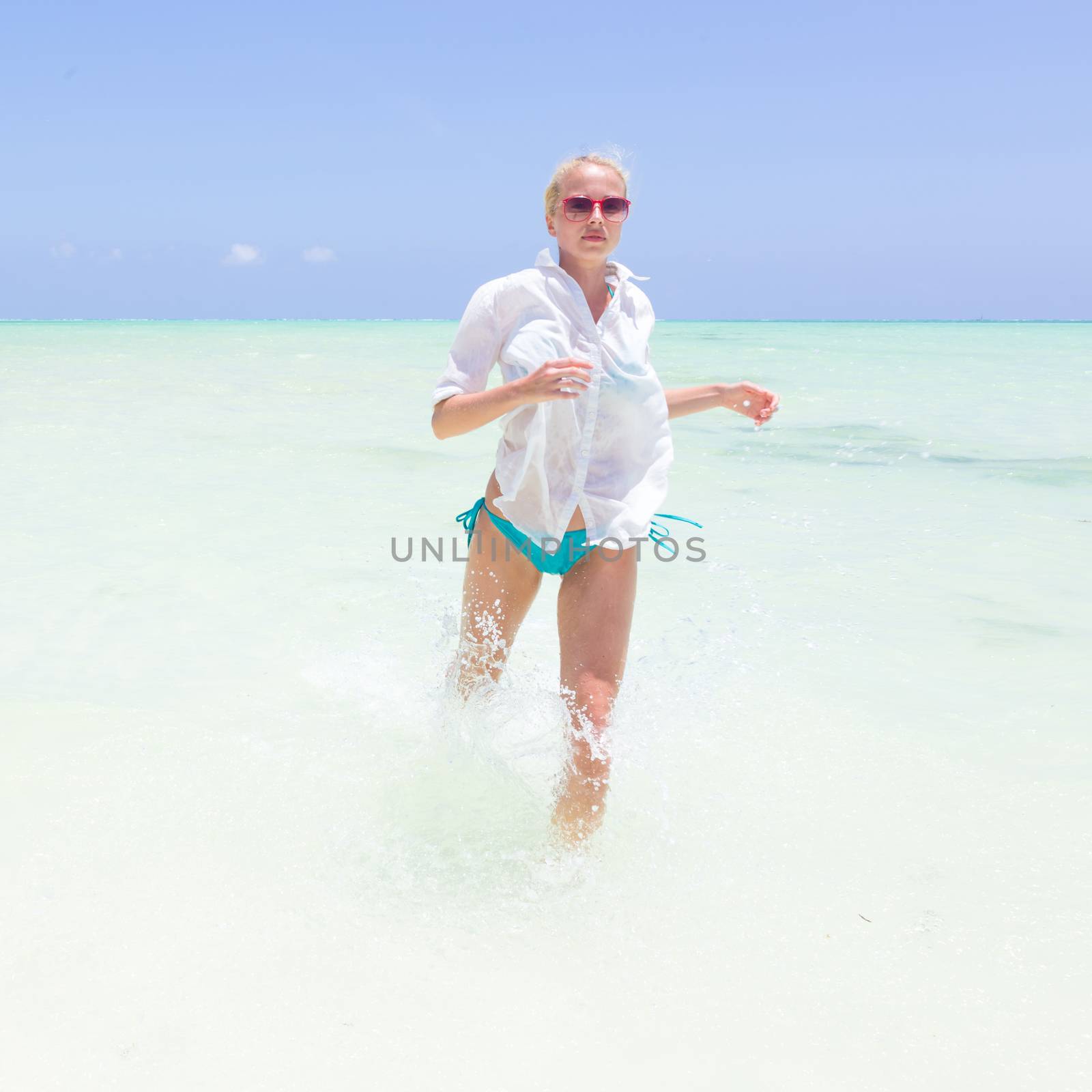 Young slim fit woman wearing white beach tunic running in sea water making water splashes with her legs. Vacation concept. Summer mood. Tropical beach setting. Paje, Zanzibar, Tanzania.