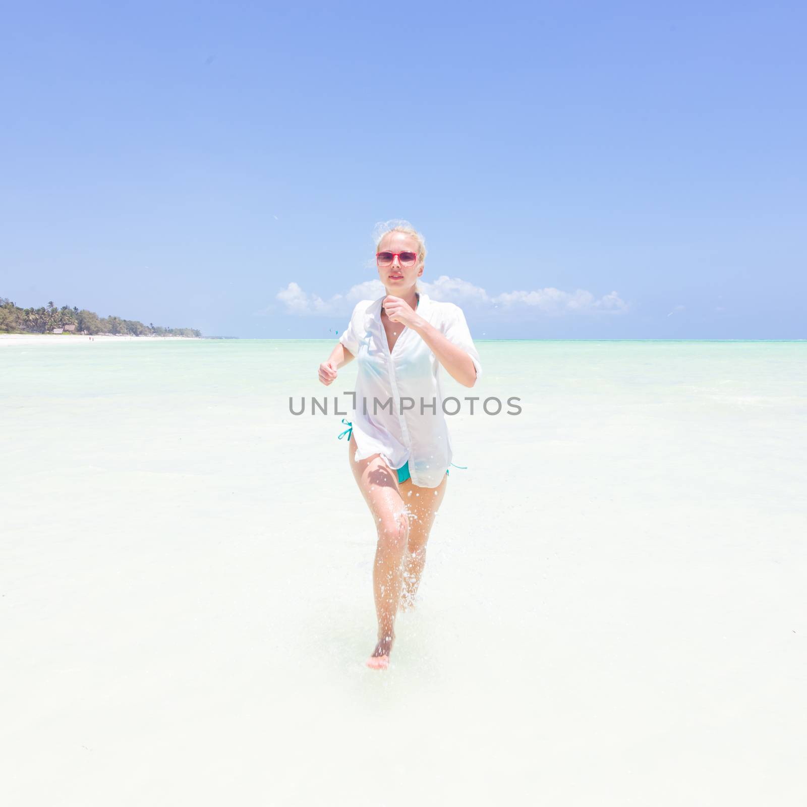 Young slim fit woman wearing white beach tunic running in sea water making water splashes with her legs. Vacation concept. Summer mood. Tropical beach setting. Paje, Zanzibar, Tanzania.