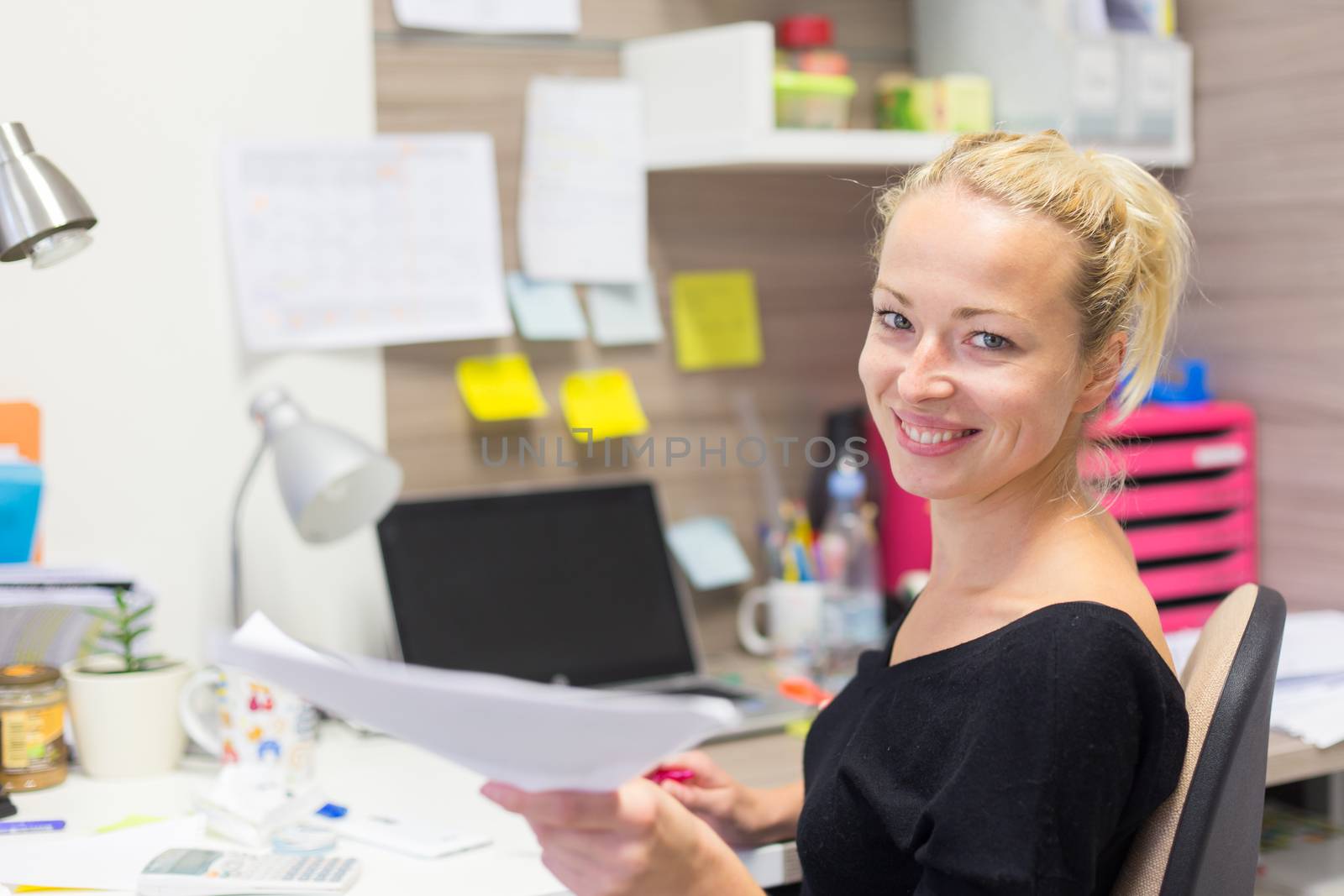 Business and entrepreneurship consept. Beautiful blonde business woman talking on mobile phone in colorful modern creative working environment accepting papers. Female multi tasking.