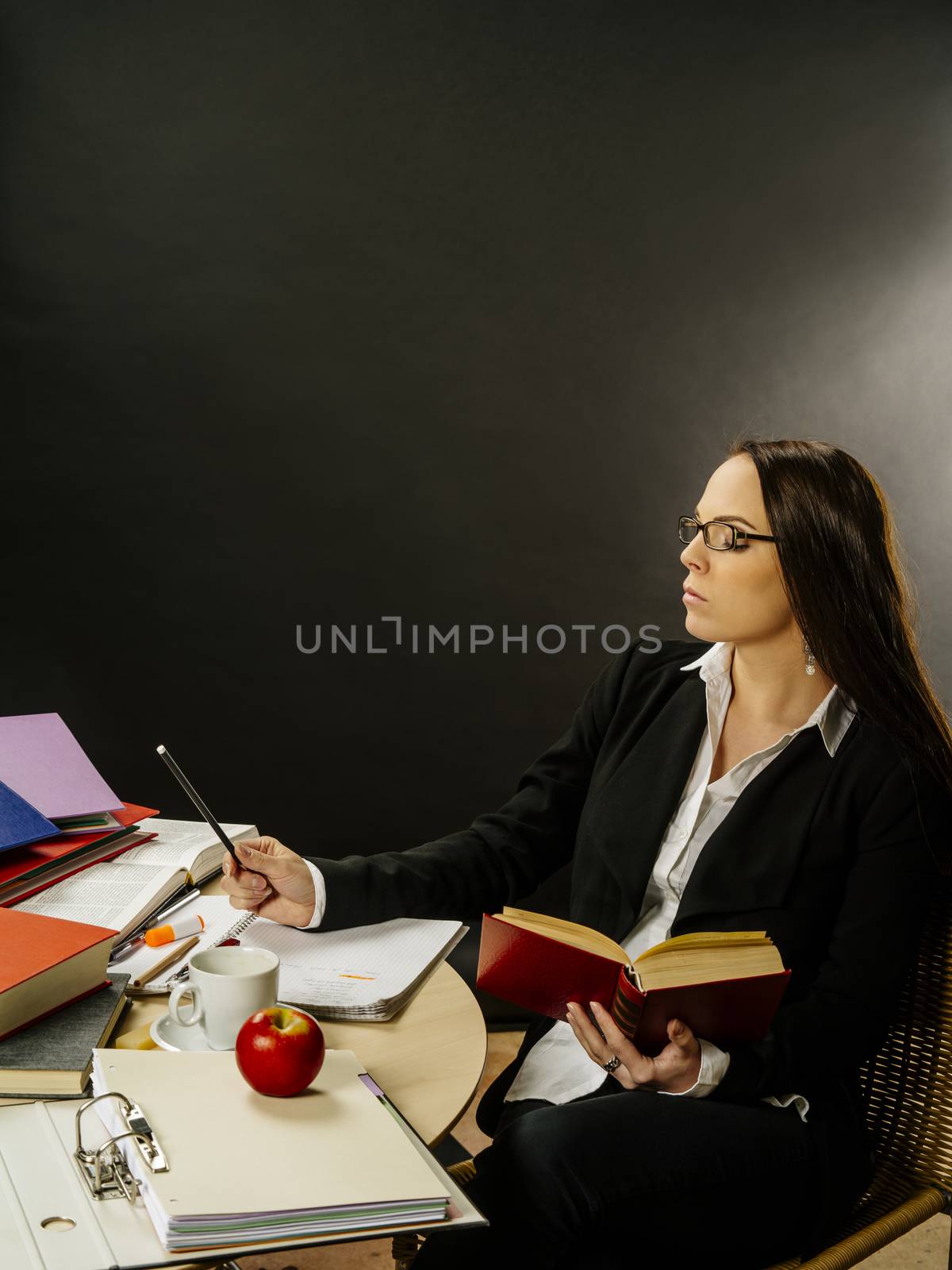 Photo of a teacher or business woman in her 30's sitting at a desk in front of a large blackboard reading and writing.