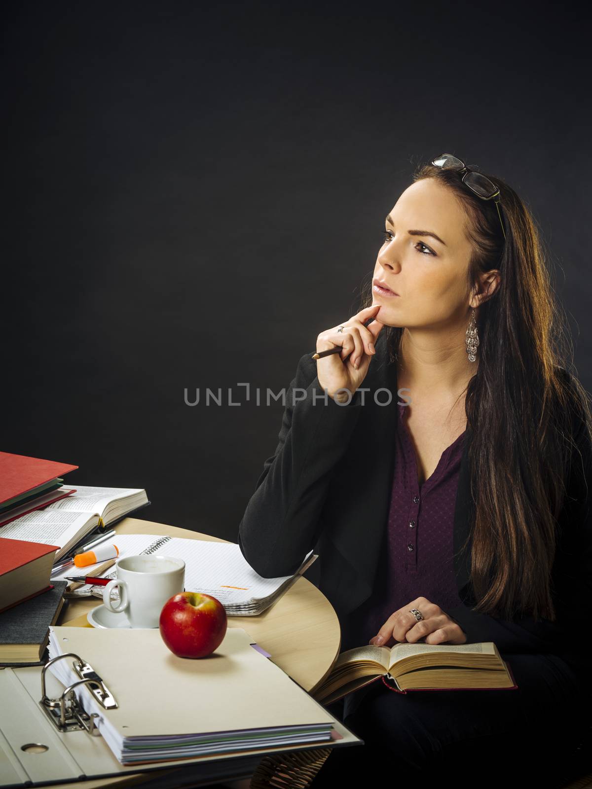 Photo of a teacher in her 30's sitting at a messy desk in front of a large blackboard reading a book.