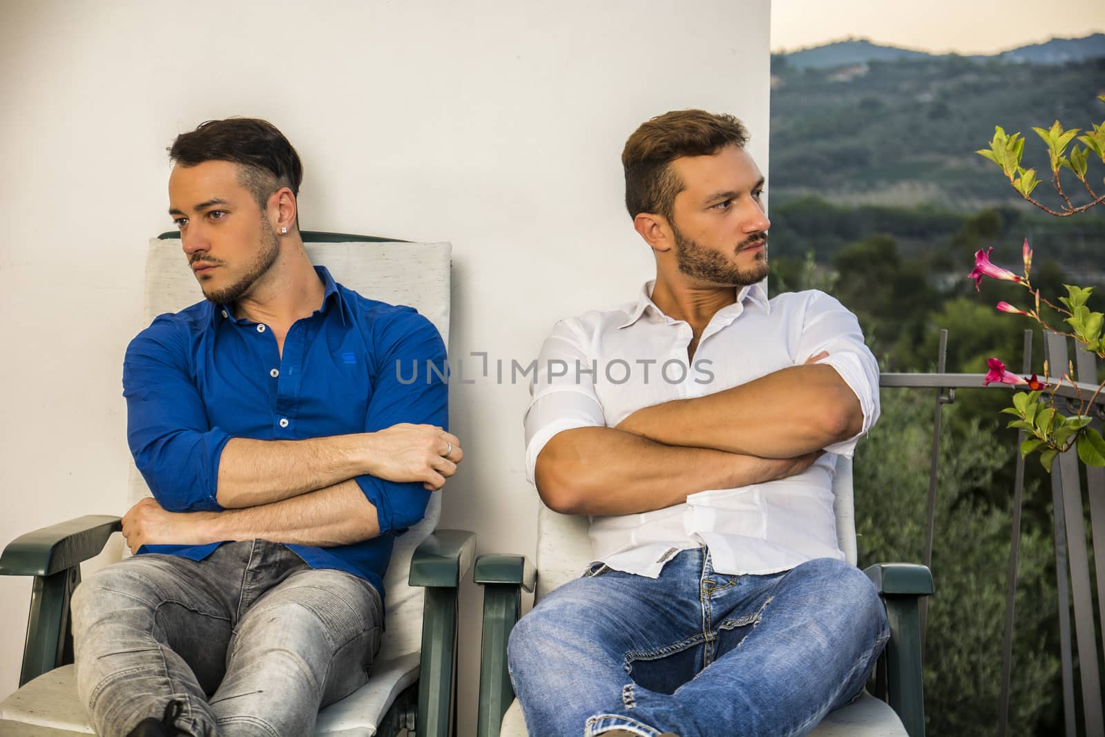 Adult gay couple in quarrel sitting in loungers with arms crossed.