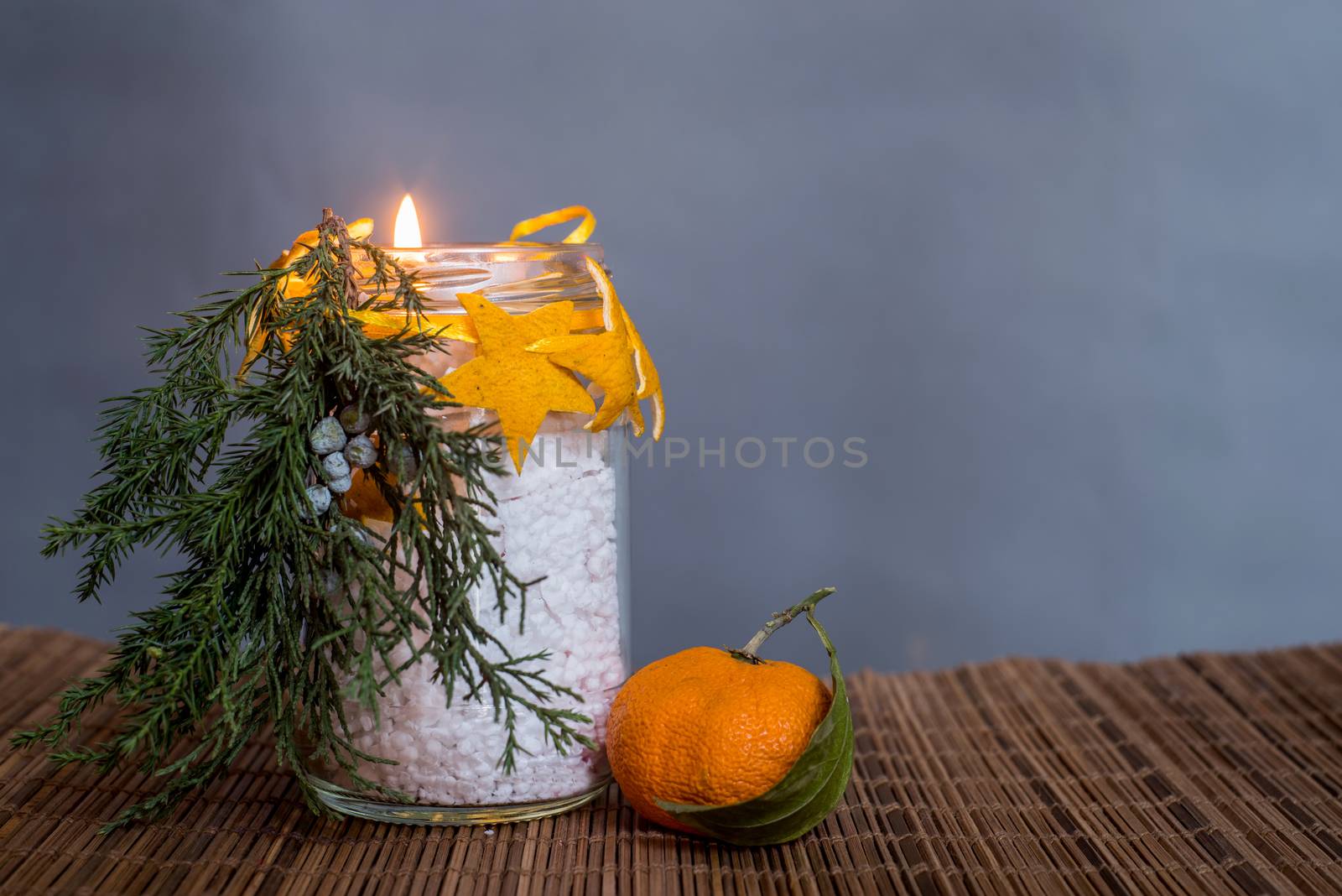 Christmas decorations on the table, hand made lit candle in a glass bottle with orange peel stars on the wooden tablecloth with a tangerine on grey