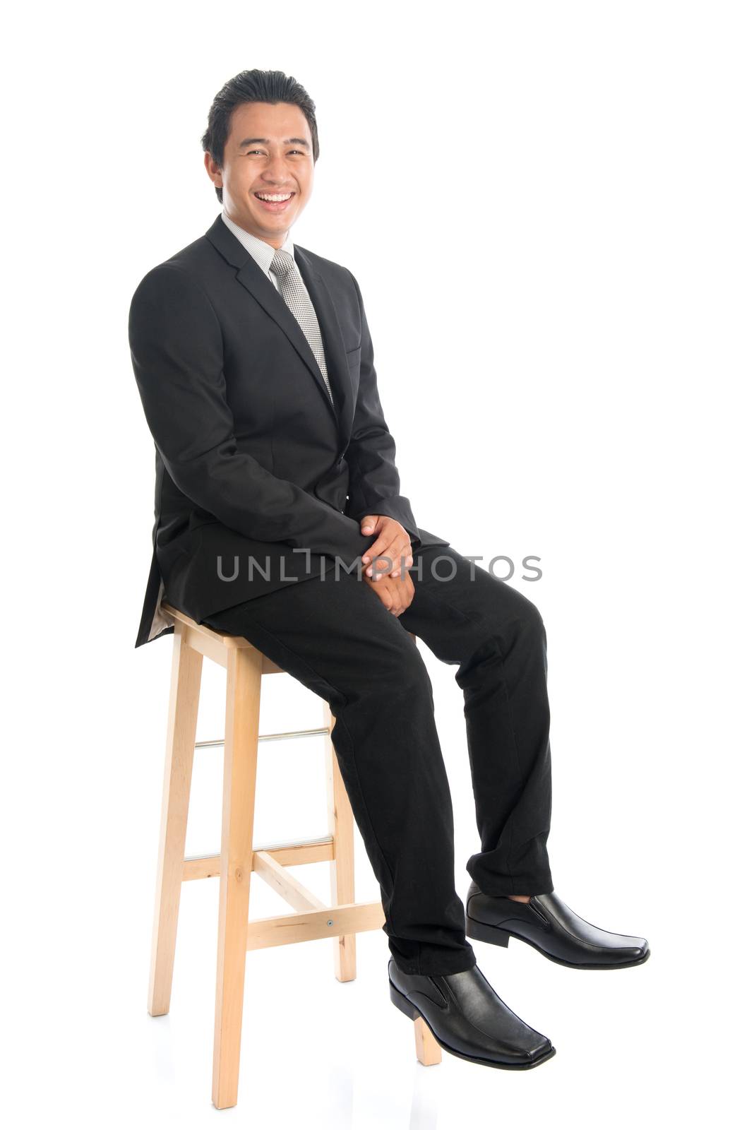 Full body portrait of handsome young Southeast Asian businessman sitting on high chair, isolated on white background. 