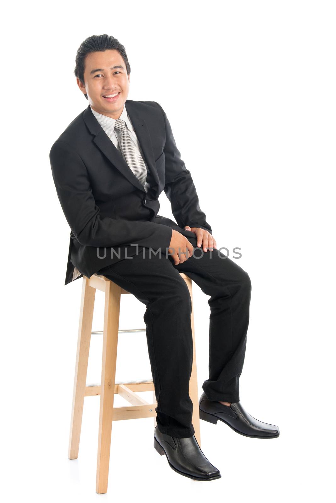 Full body portrait of attractive young Southeast Asian businessman sitting on high chair, isolated on white background. 