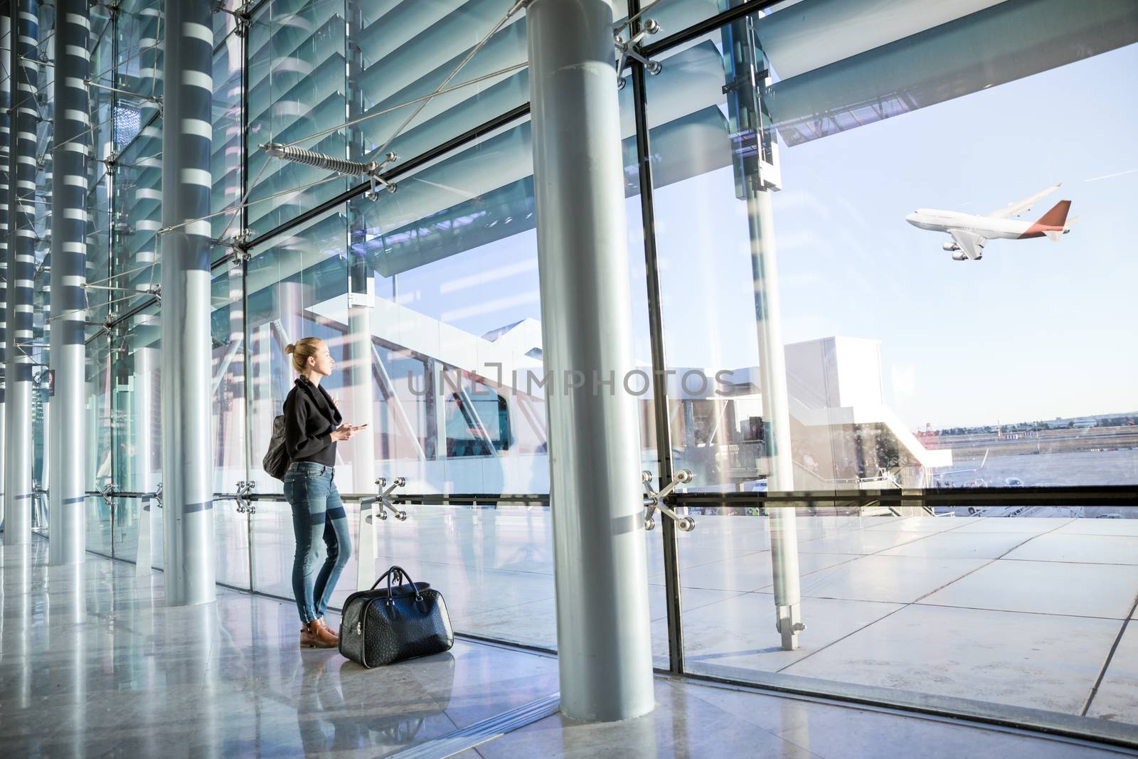Young casual female traveler at airport, holding smart phone device, looking through the airport gate windows at planes on airport runway.