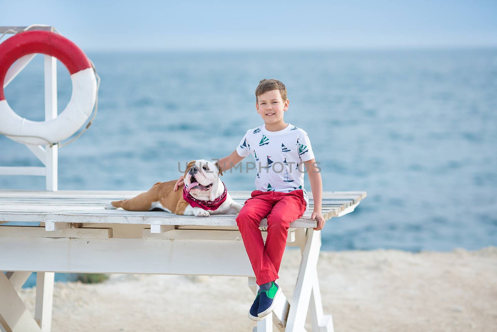 Handsome boy teen happyly spending time together with his friend bulldog on sea side Kid dog holding playing two sea stars close to life buoy float wearing red pants trousers slippers and t-shirt.