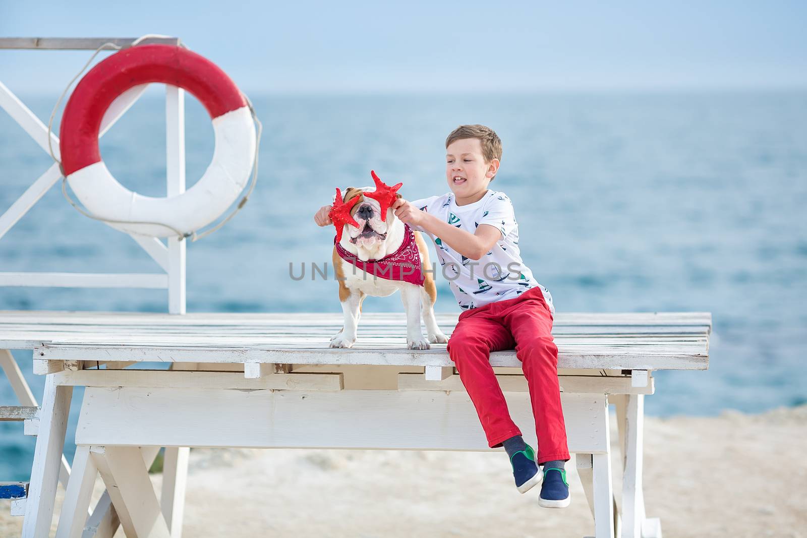 Handsome boy teen happyly spending time together with his friend bulldog on sea side Kid dog holding playing two sea stars close to life buoy float wearing red pants trousers slippers and t-shirt by dlukashenko@mail.ru