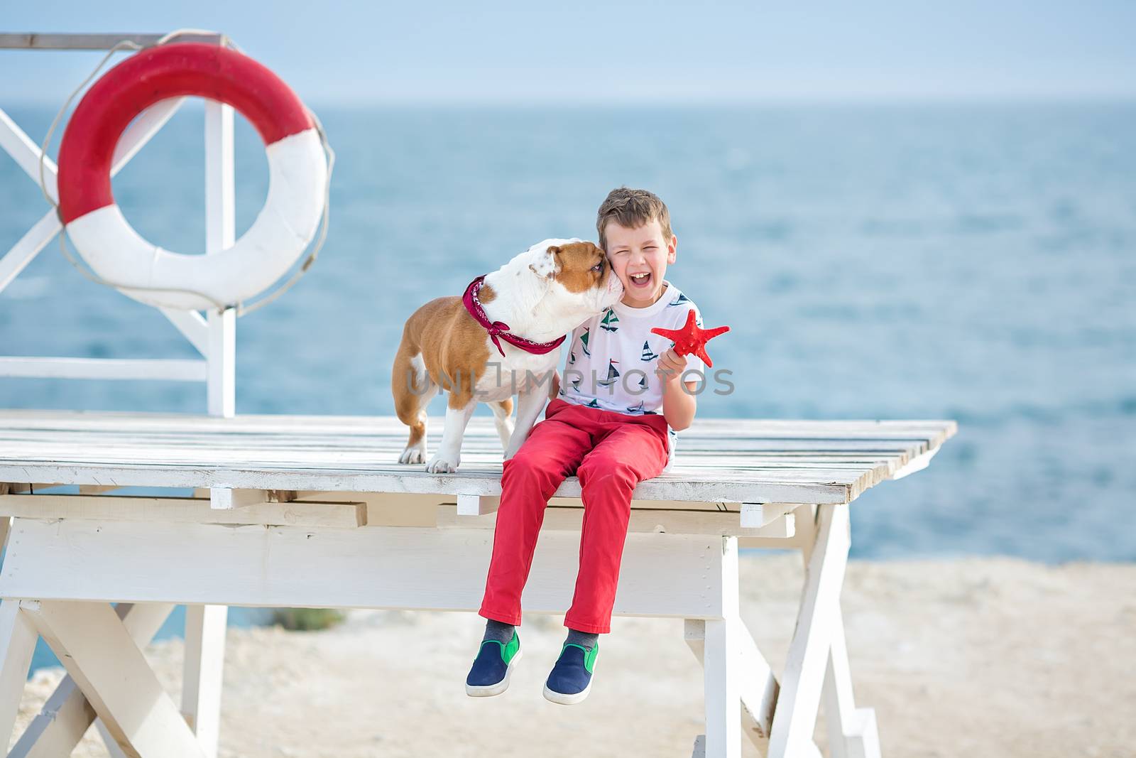 Handsome boy teen happyly spending time together with his friend bulldog on sea side Kid dog holding playing two sea stars close to life buoy float wearing red pants trousers slippers and t-shirt.