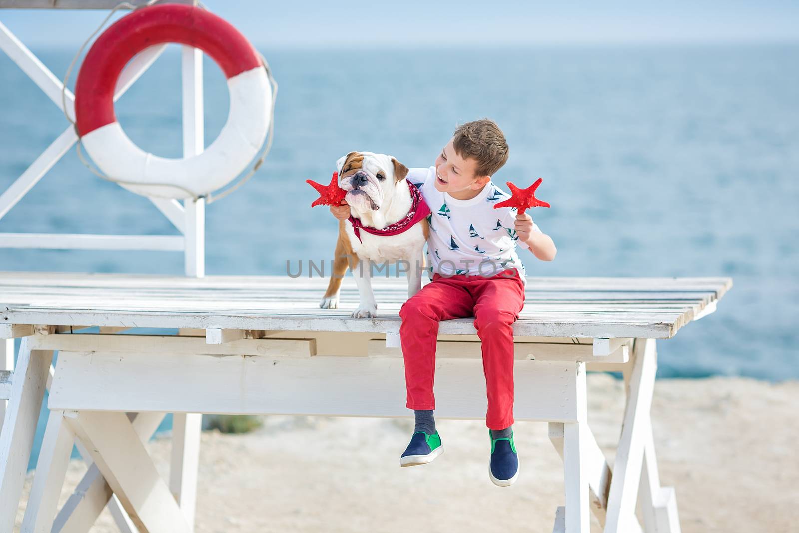 Handsome boy teen happyly spending time together with his friend bulldog on sea side Kid dog holding playing two sea stars close to life buoy float wearing red pants trousers slippers and t-shirt by dlukashenko@mail.ru