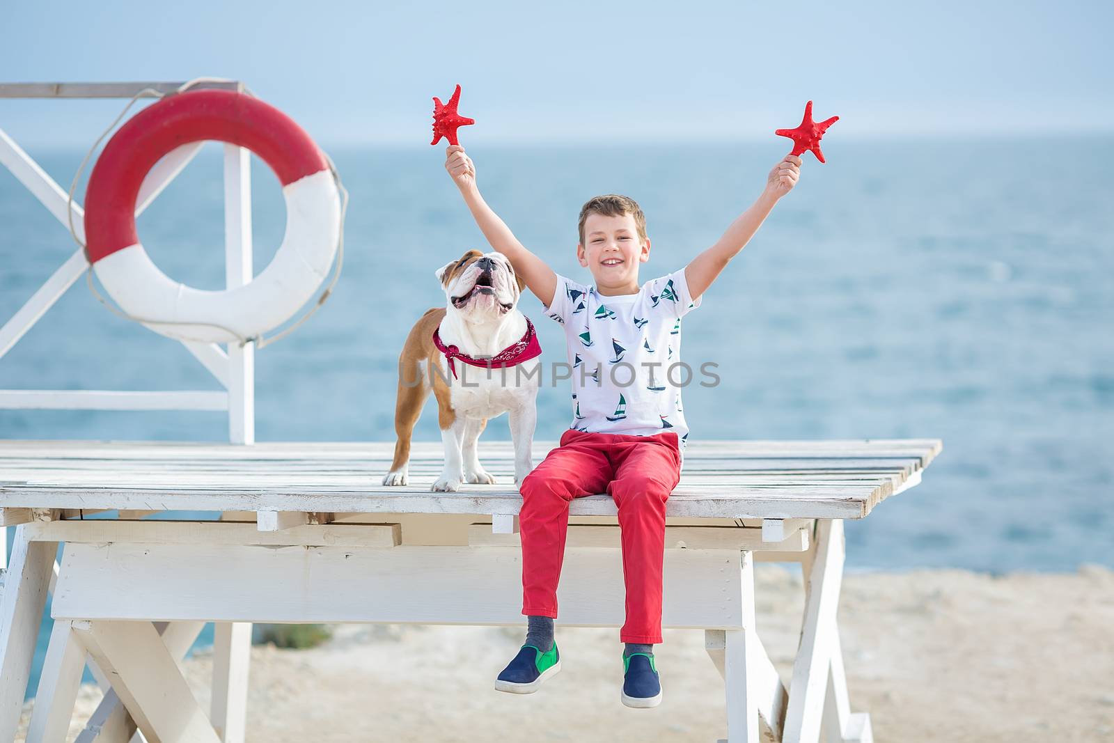 Handsome boy teen happyly spending time together with his friend bulldog on sea side Kid dog holding playing two sea stars close to life buoy float wearing red pants trousers slippers and t-shirt by dlukashenko@mail.ru