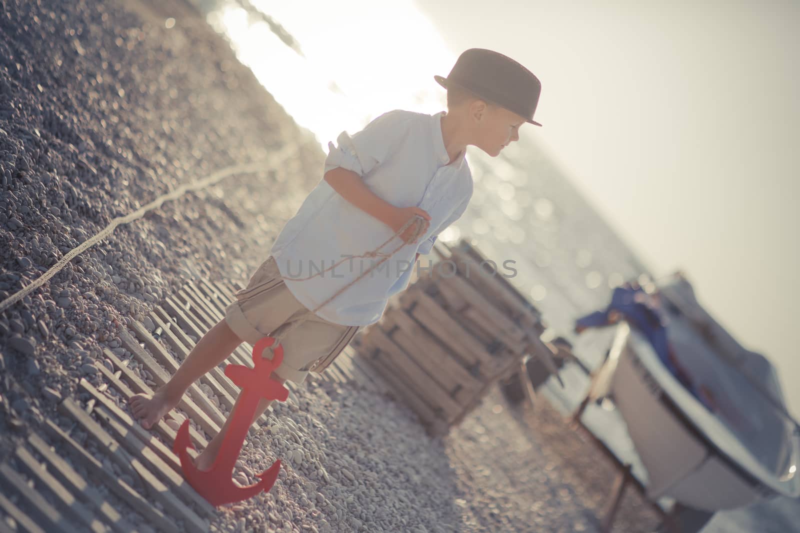Handsome Nice looking boy on beach walking posing on wooden way wearing fancy stylish blue shirt and shorts with gallows and gentleman hat enjoy summer time alone on awesome ocean with anchor.