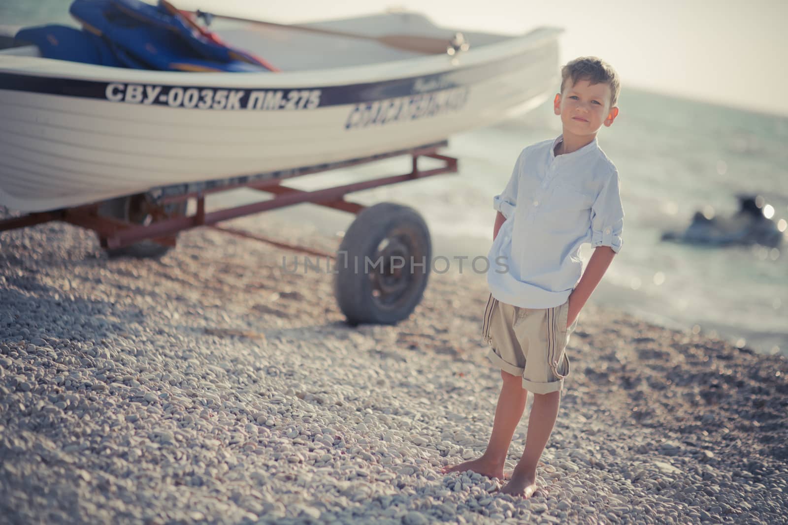 Nice looking handsome boy on beach close to wooden boat wearing fancy stylish blue shirt and shorts with gallows enjoy summer time alone on awesome ocean.