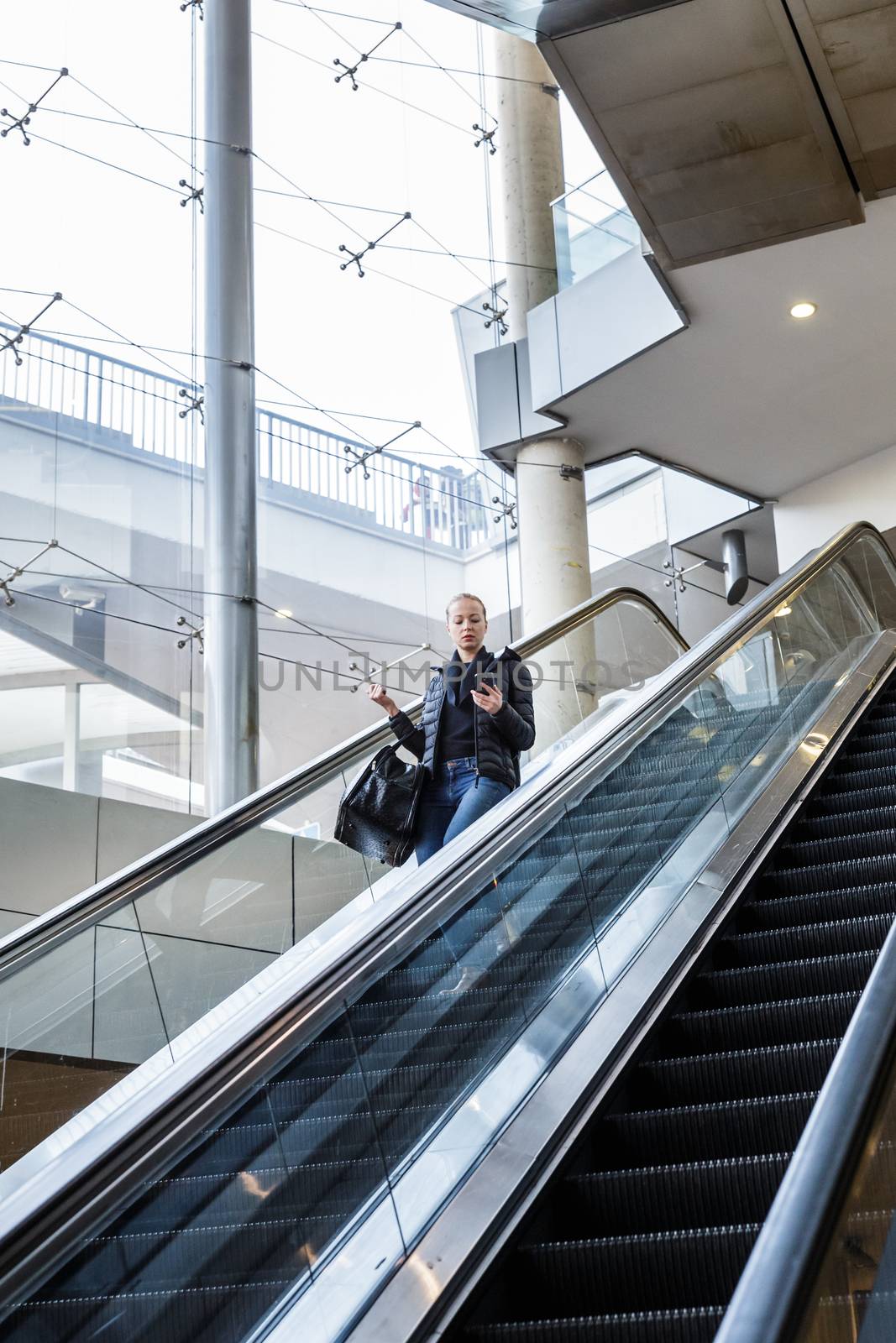 Young businesswoman with large black bag and mobile phone getting down on the escalator during business trip in modern city.