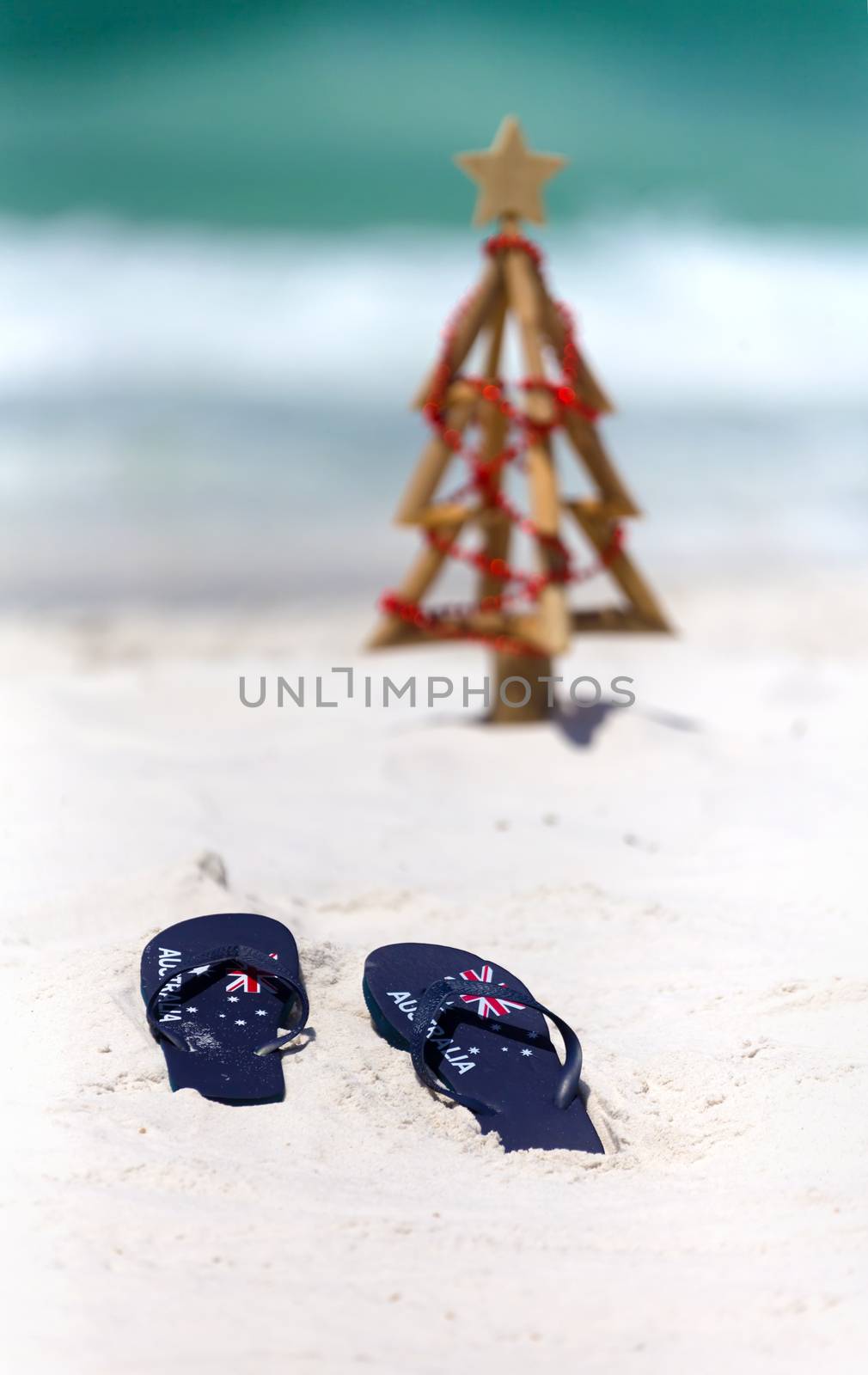 Australia flag thongs in focus with a small diriftwood Christmas tree and ocean waves in the background.