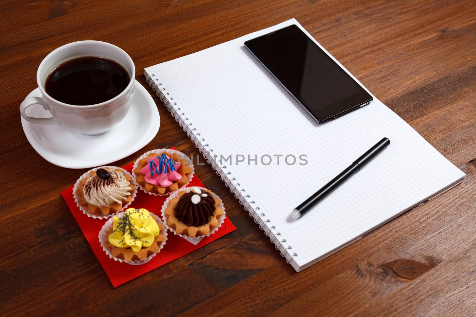 White coffee cup with notebook, four cupcakes, smartphone and pen on a wooden table.