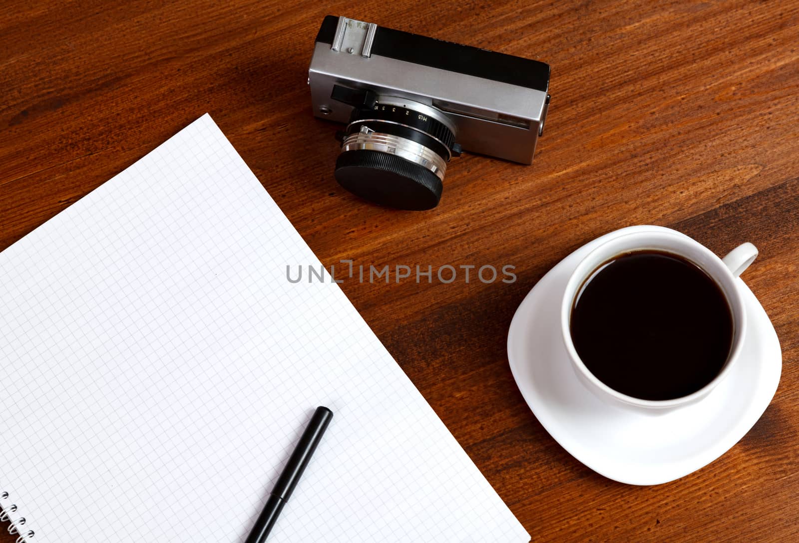 Retro film photo camera, cup of coffee and notebook with pencil on brown table. Top view.