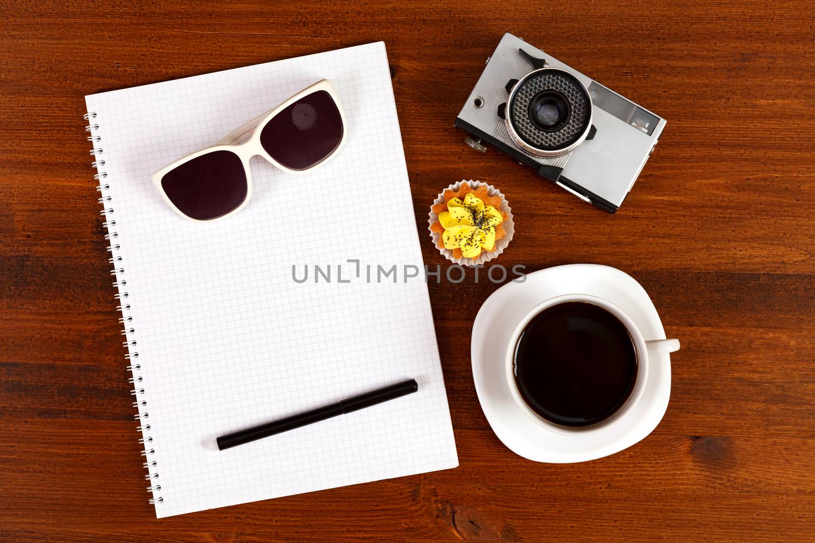 Retro film photo camera, sunglasses, cupcake, cup of coffee and notebook with pencil on brown table. Top view.