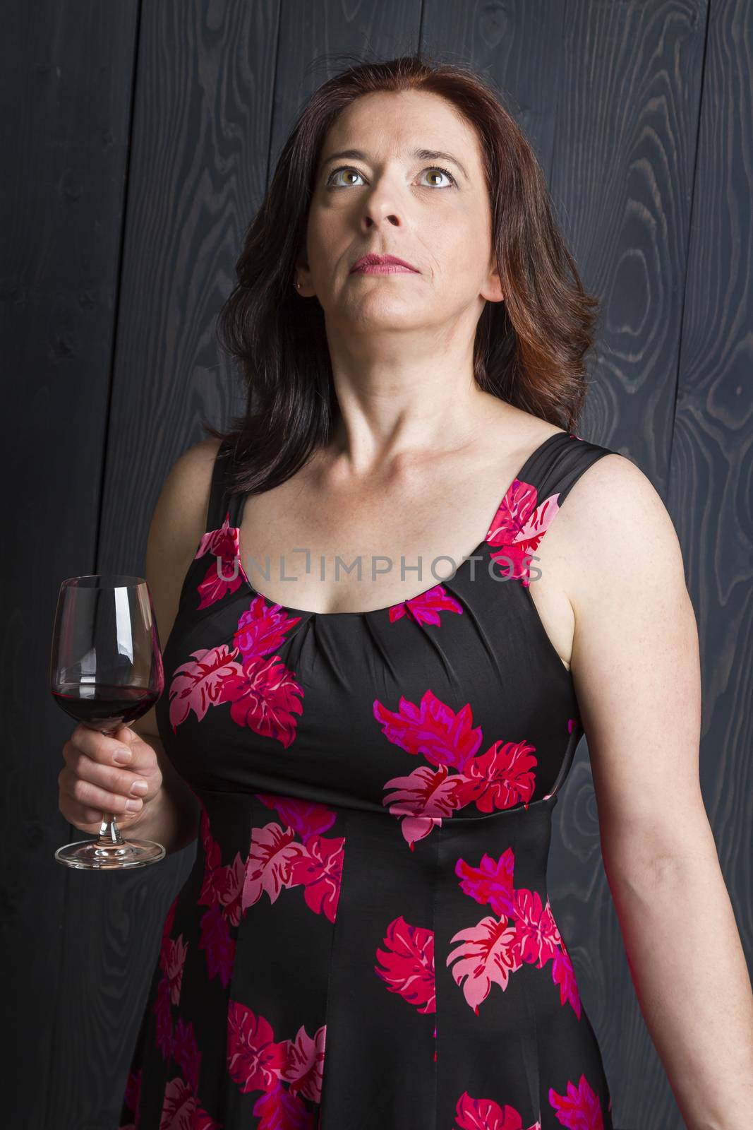 forty something brunette woman, wearing a summer dress with a glass of red wine, looking up, in front of a blue wood stain background