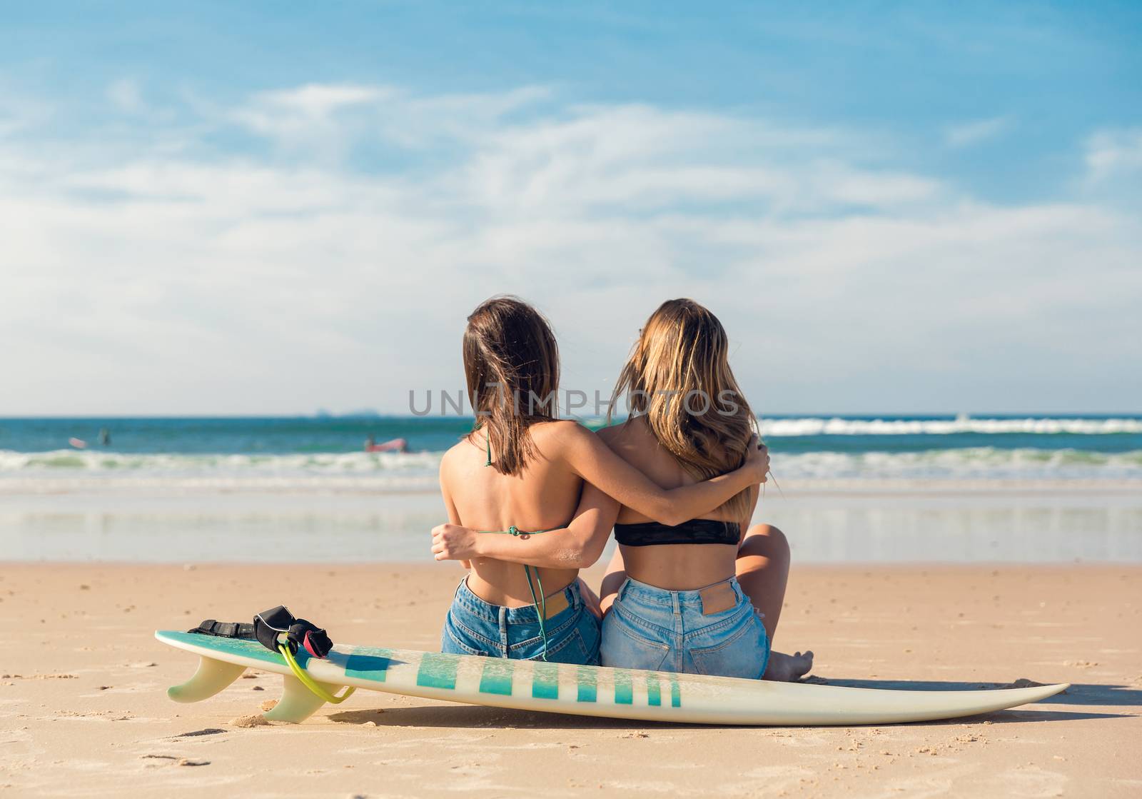 Two beautiful female friends at the beach sitting on the sand close to her surfboards embracing each other