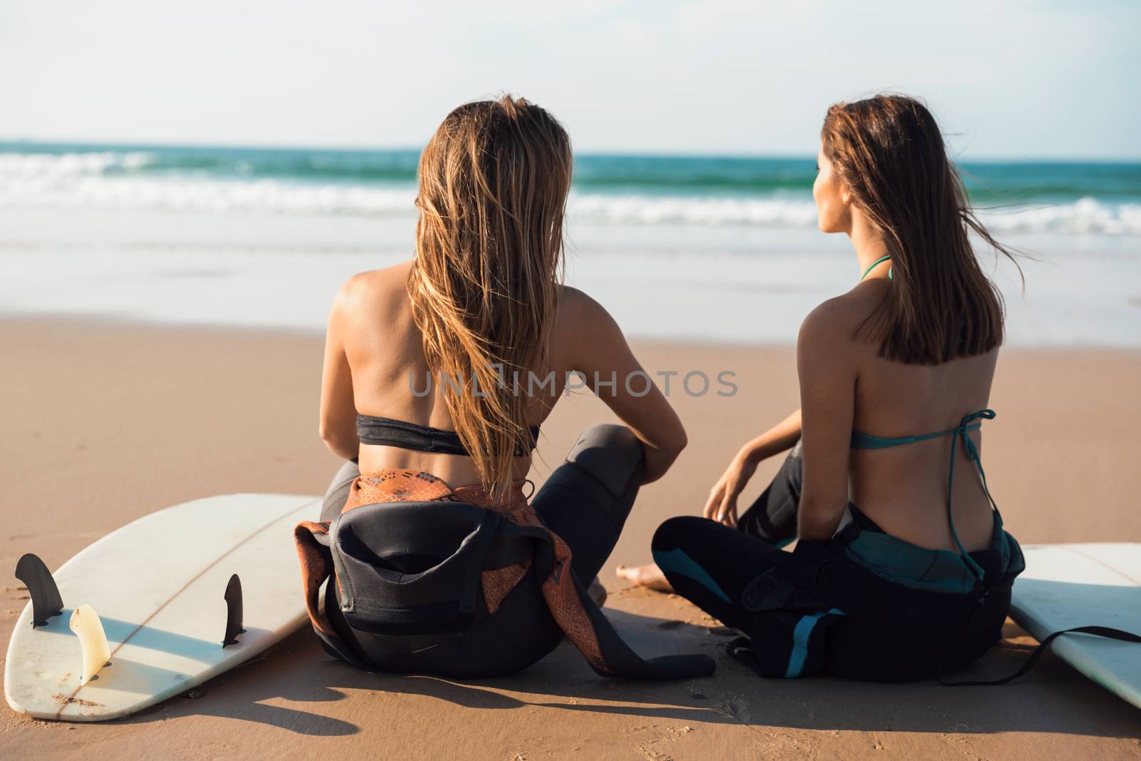 Two beautiful female friends at the beach sitting on the sand with her surfboards while looking to the ocean