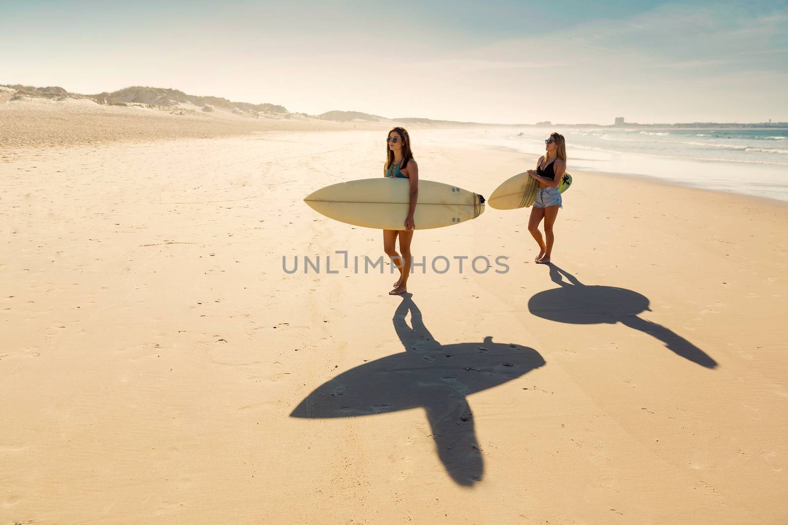 Two beautiful friends holding their surfboards and walking on the beach