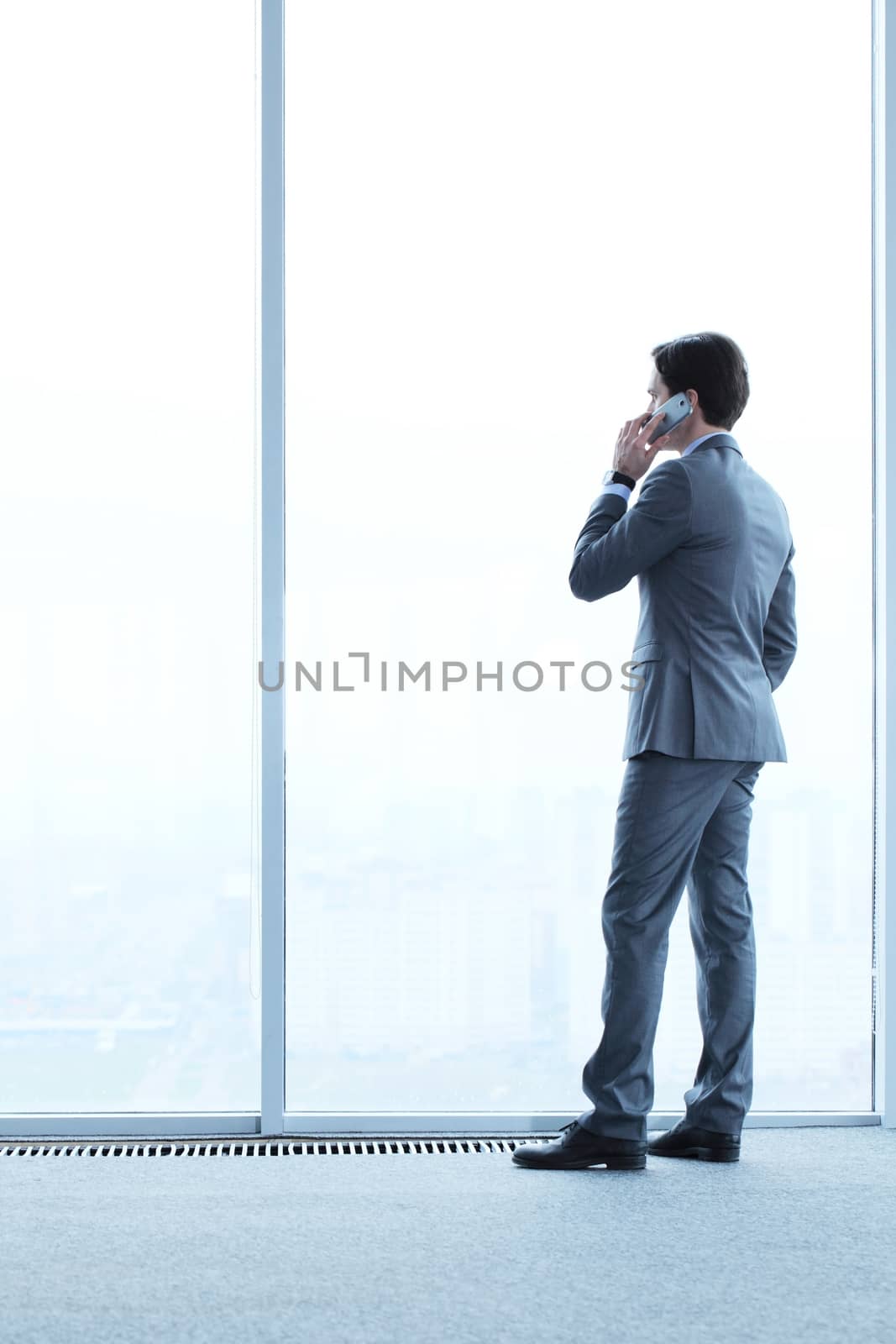 Businessman standing near window, looking at city and talking by phone