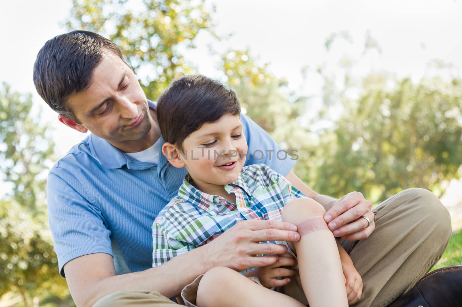 Loving Father Puts a Bandage on the Elbow of His Young Son in the Park. by Feverpitched