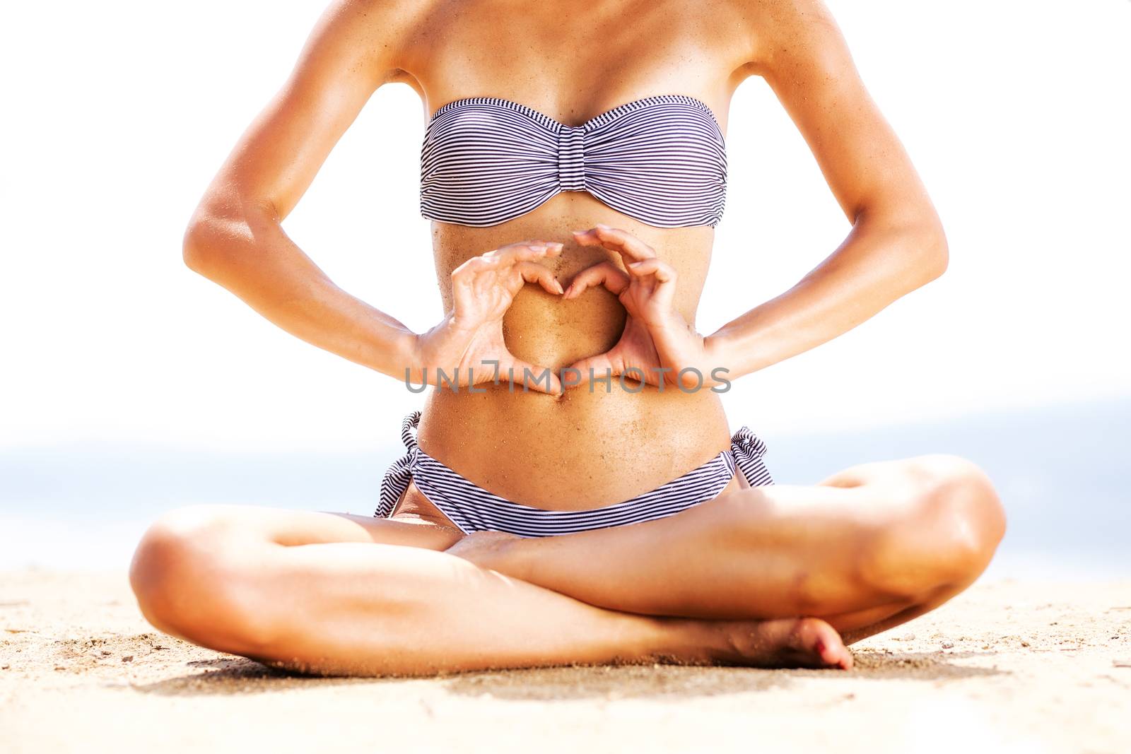 sexy female body on a beach, with sand on the belly, making a heart with her hands