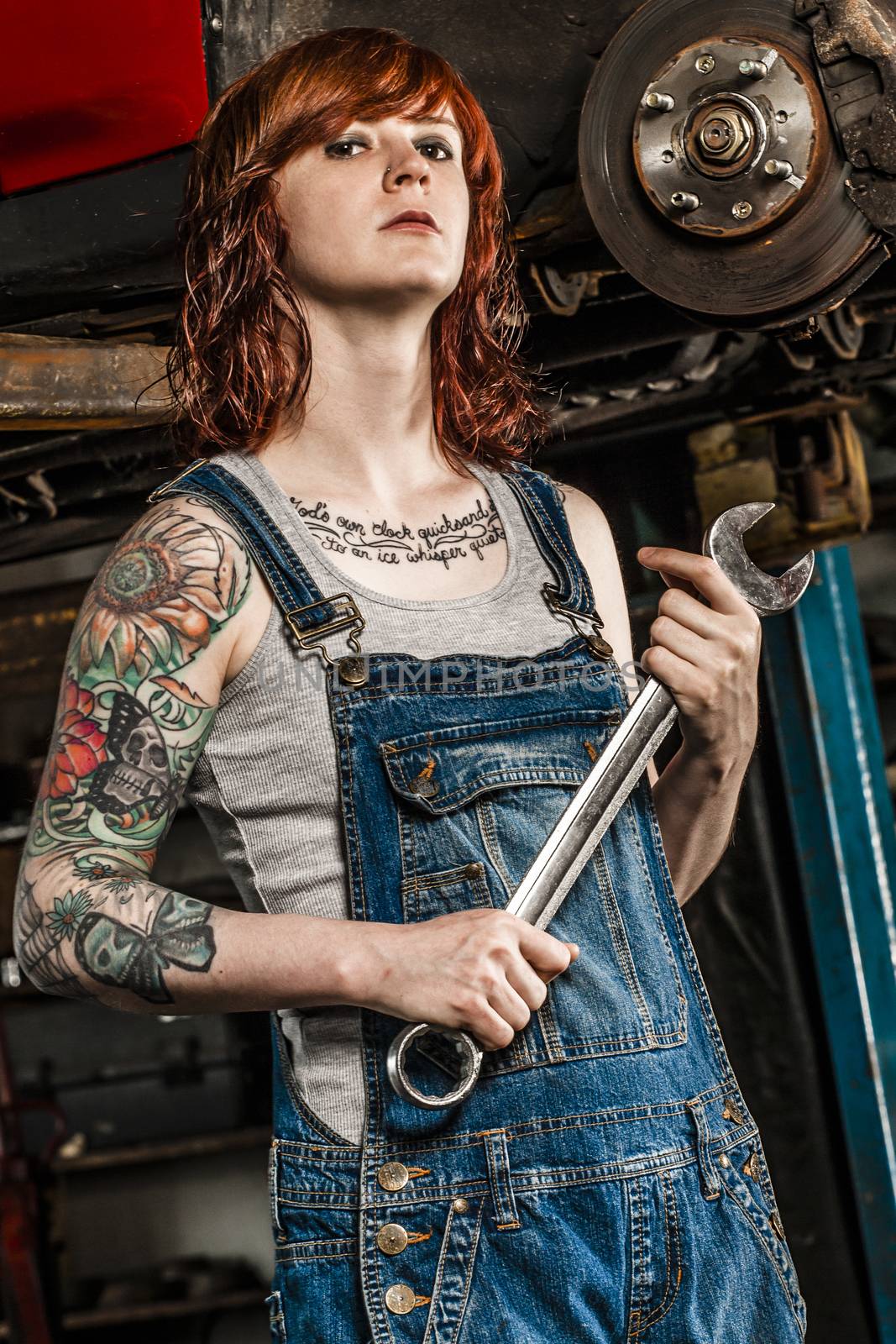 Photo of a young beautiful redhead mechanic with tattoos and wearing overalls fixing the brakes on a car. Attached property release is for arm tattoos.