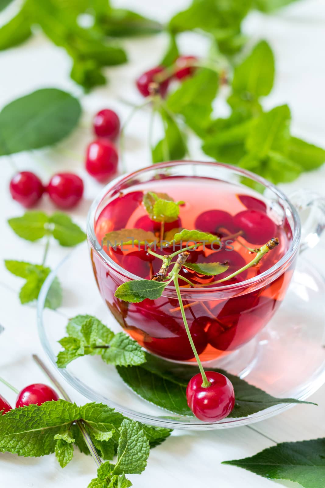 Fresh fruit cherry drink in transparent glass cup surrounded by cherries on the white wooden background.